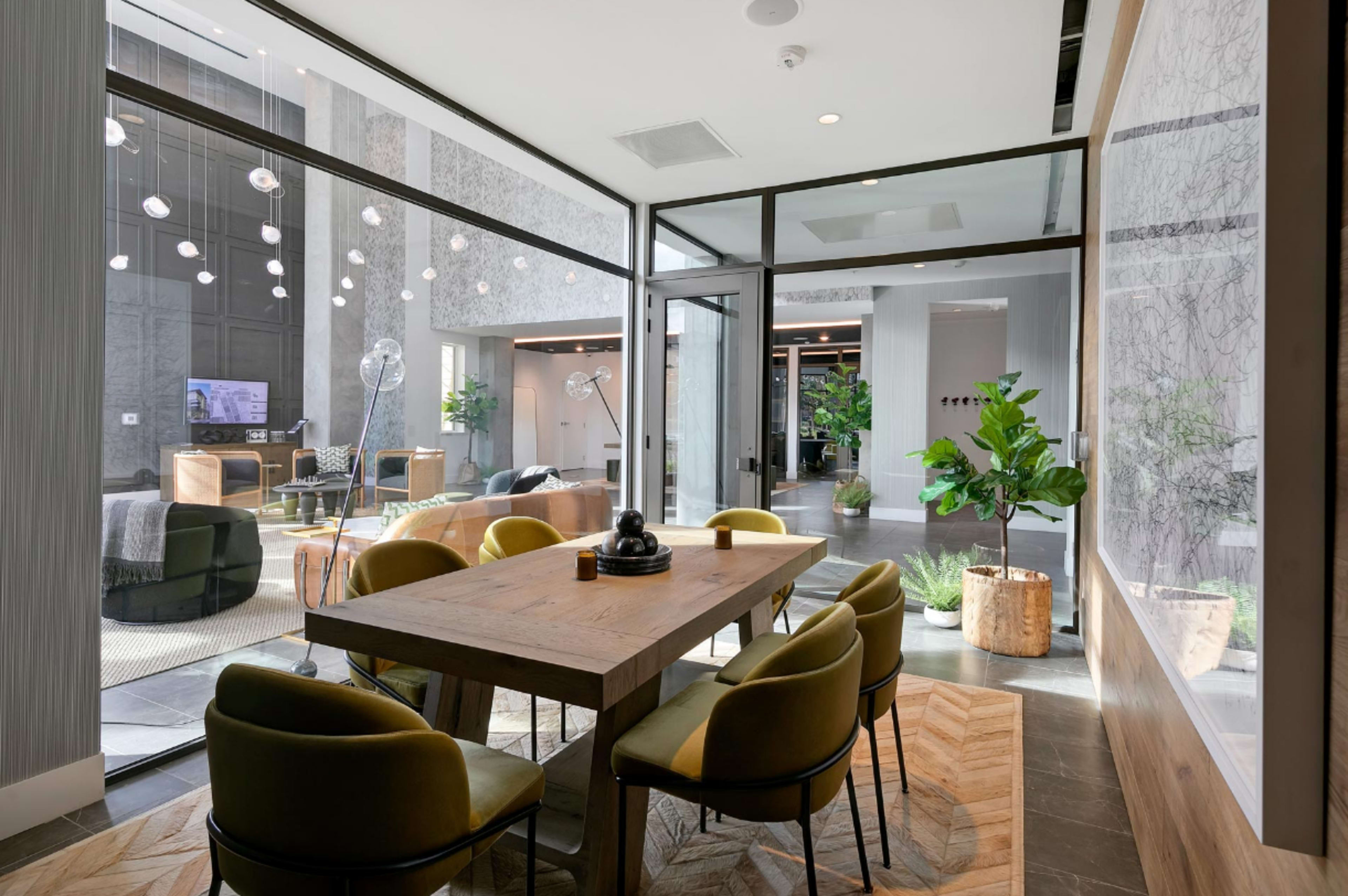 A modern office space features a large wooden table surrounded by green chairs, with a view of a lounge area and large glass windows allowing natural light.
