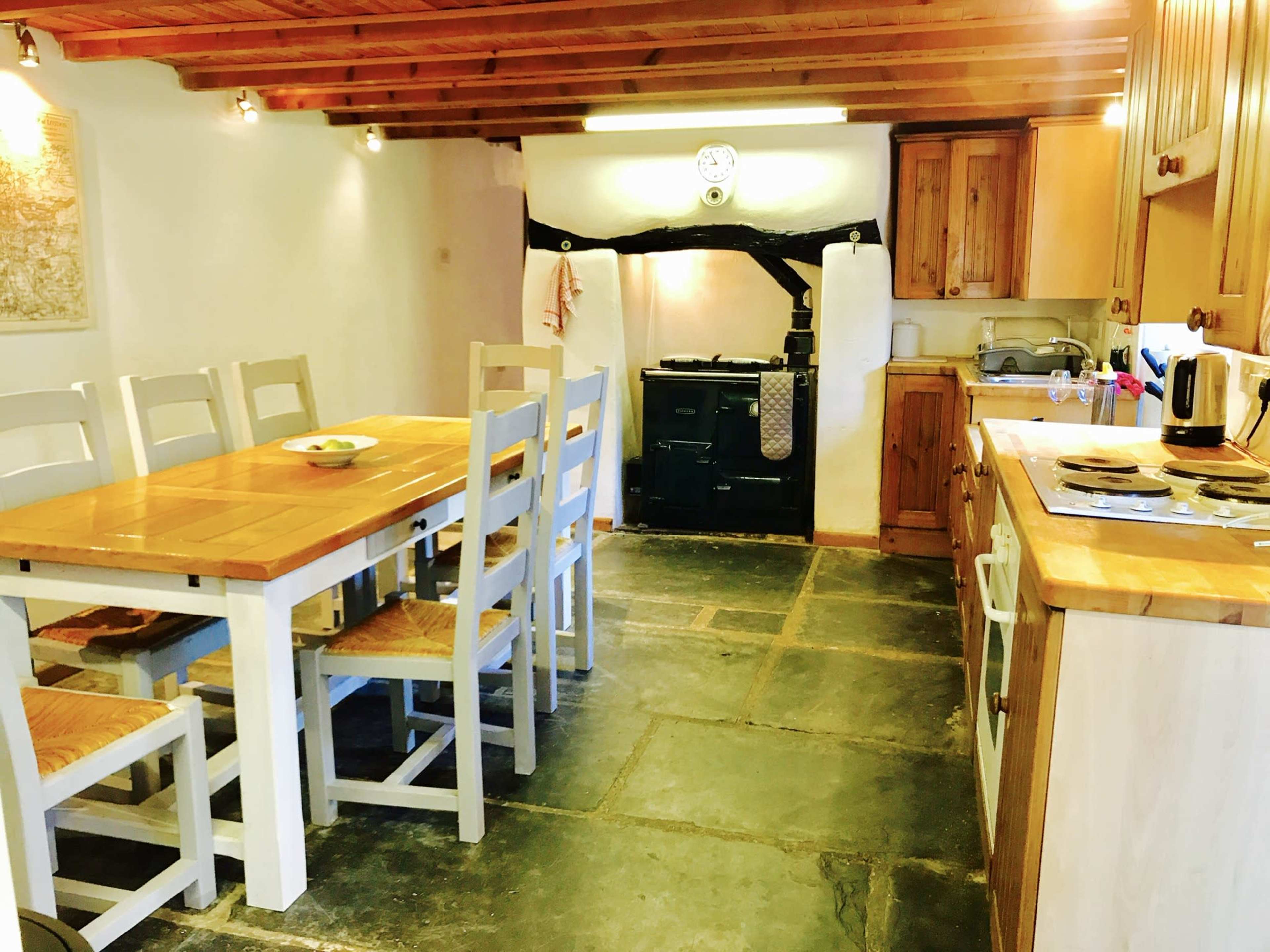 A rustic kitchen features a large wooden table with chairs, an Aga stove, and various wooden cabinets along the walls.