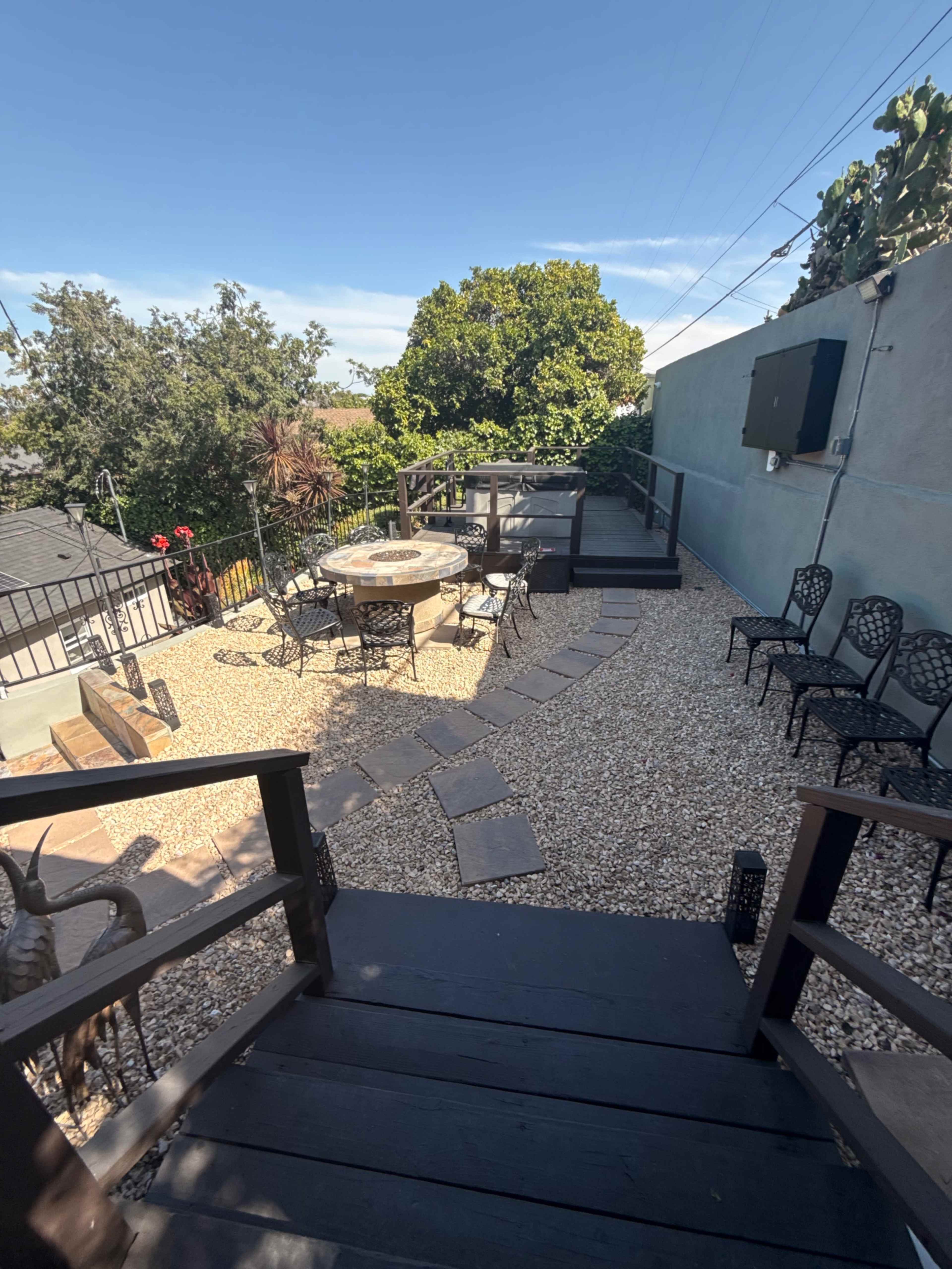 The image shows a landscaped outdoor area with a stone pathway, a round table surrounded by benches, and several metal chairs on a gravel surface.