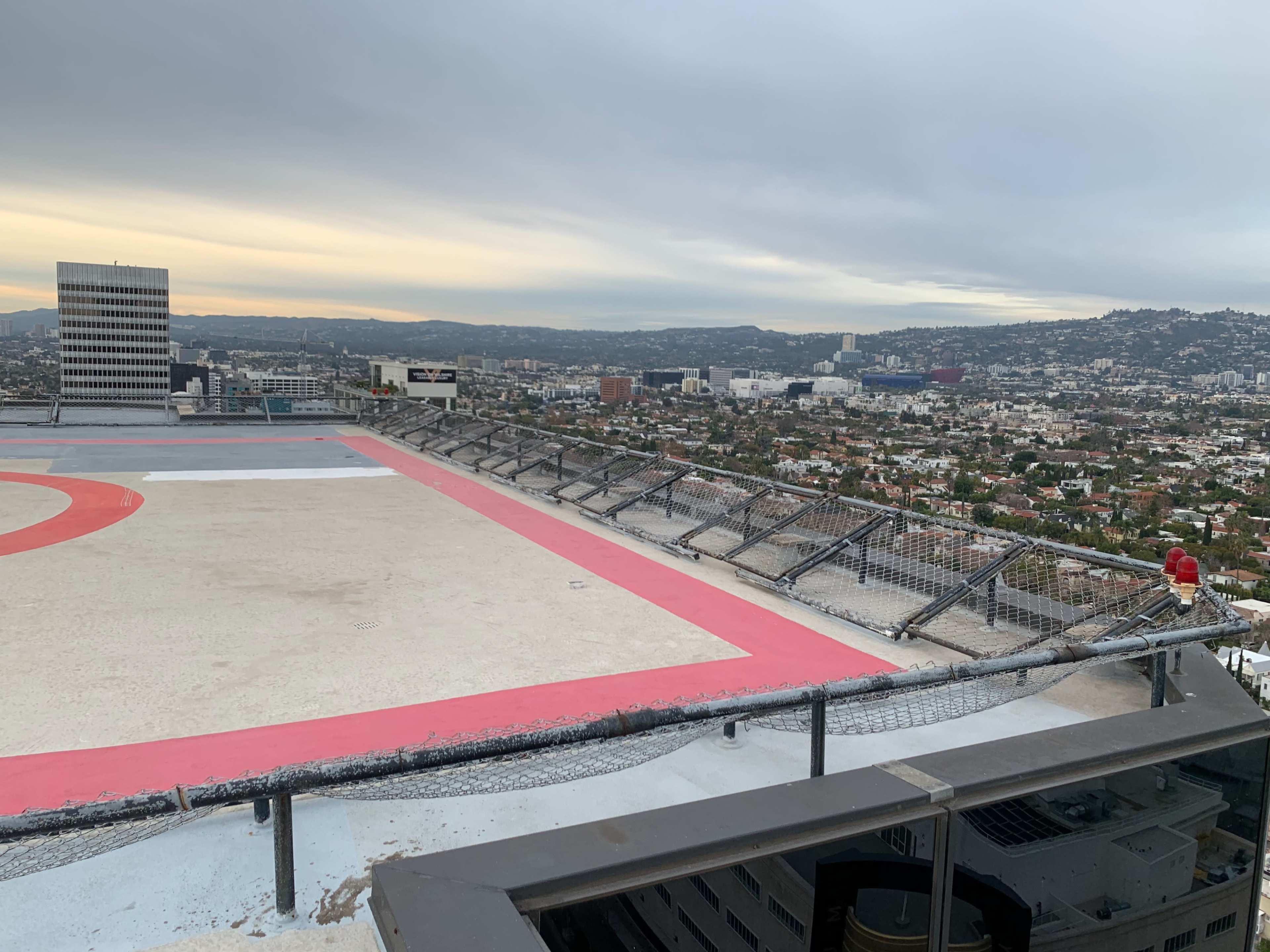 A helipad is positioned on the rooftop of a building overlooking a cityscape with distant mountains and overcast skies.