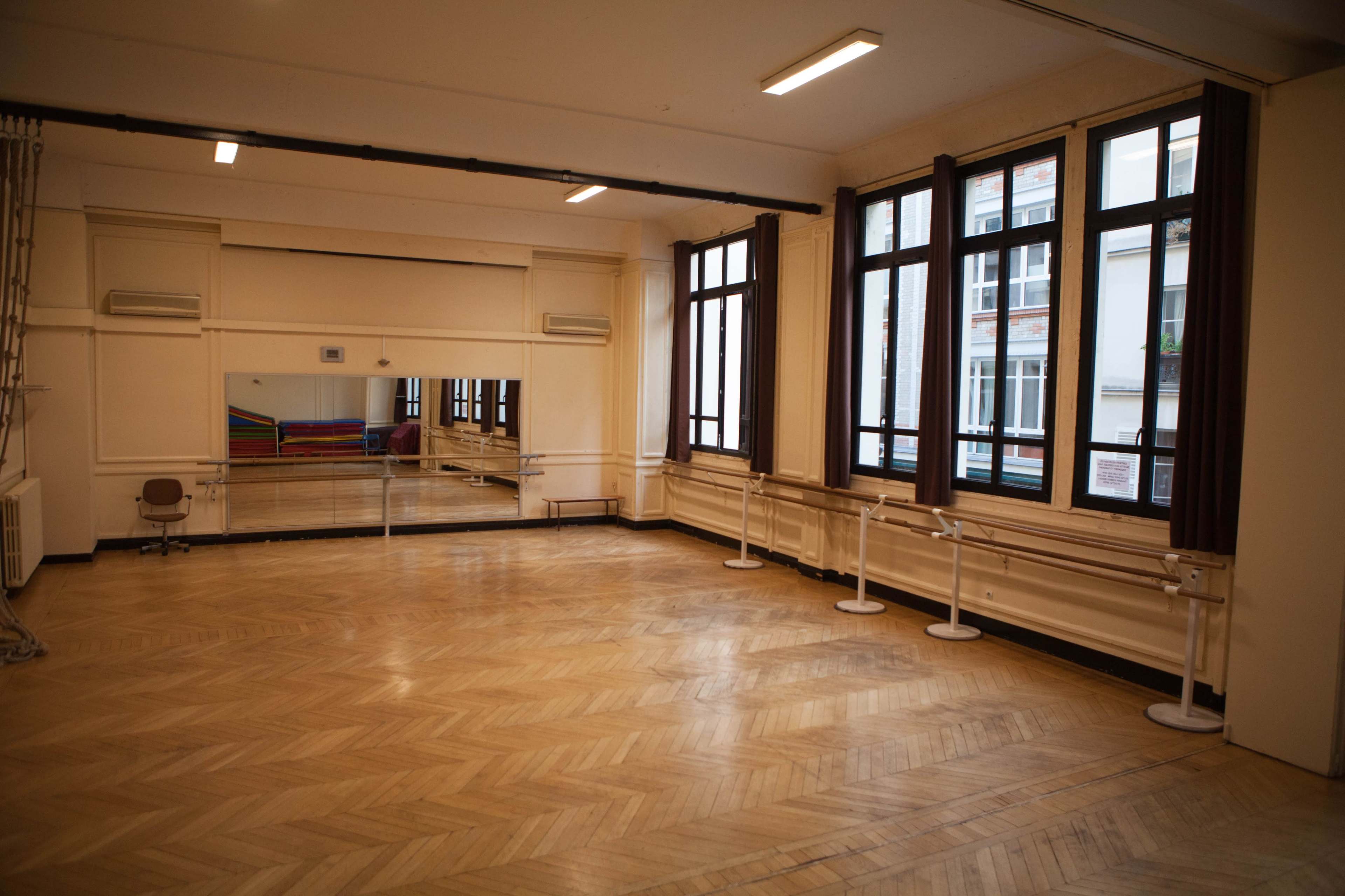 The image shows an empty dance studio with wooden flooring, large windows, and a wall mirror.
