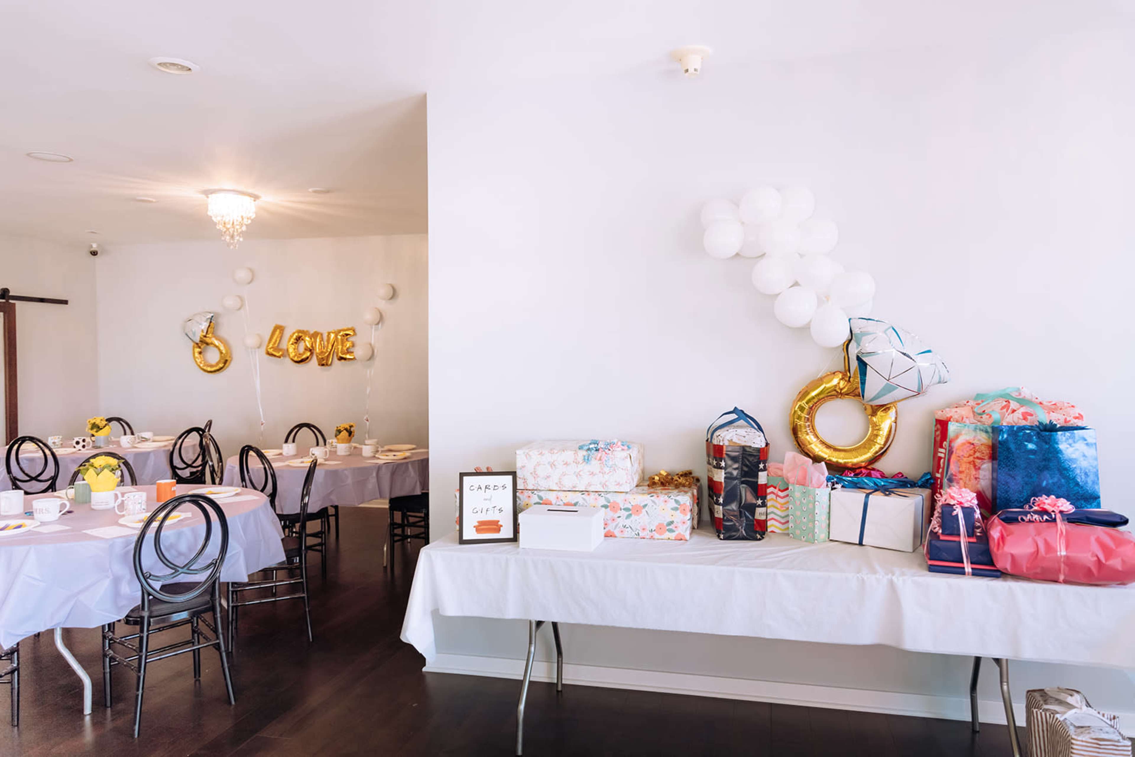 A dining area features tables set with white tablecloths and a gift table adorned with colorful wrapped presents and balloons.
