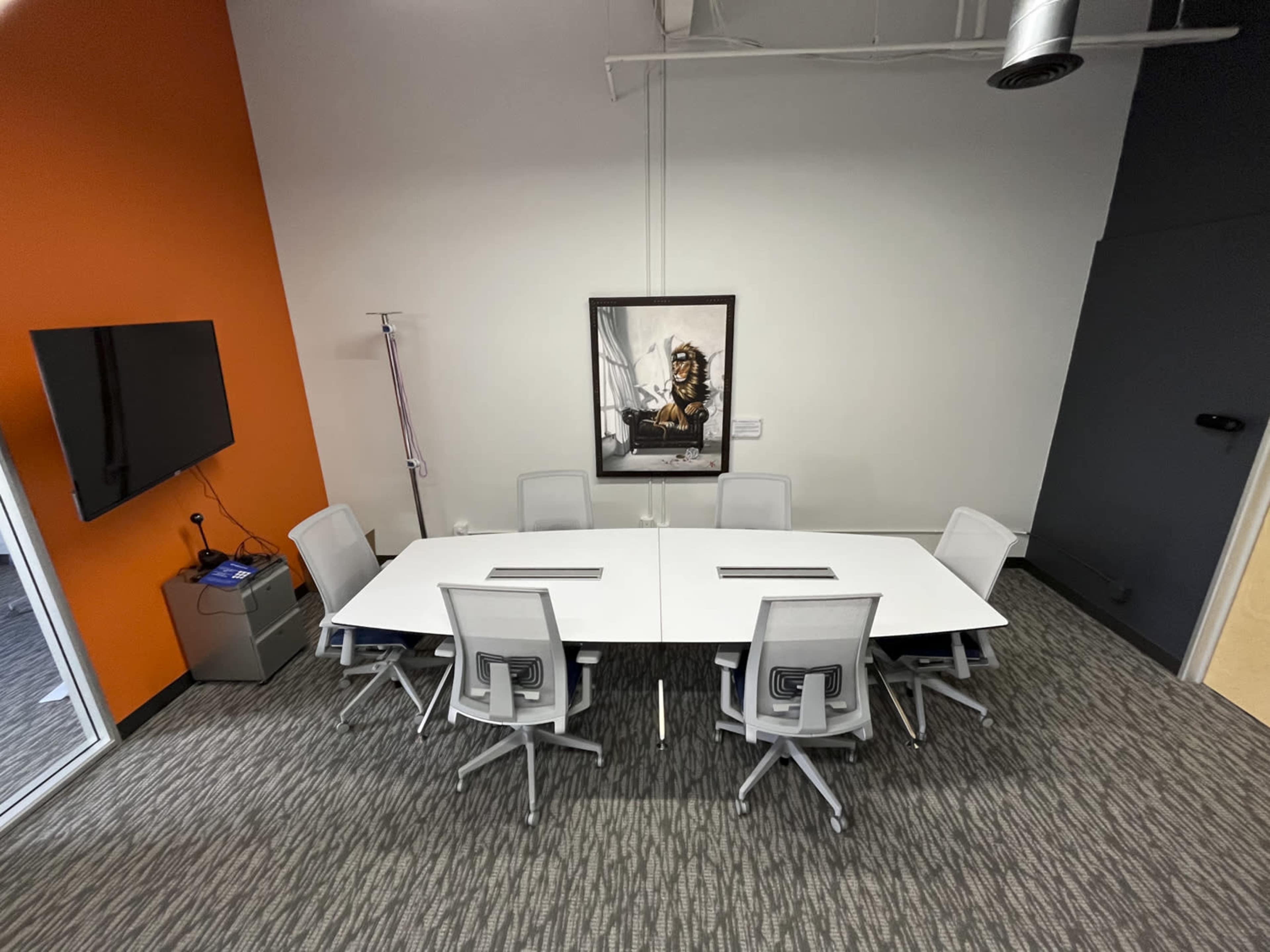 A modern conference room features a large white table surrounded by grey chairs, with an orange wall and a framed artwork.