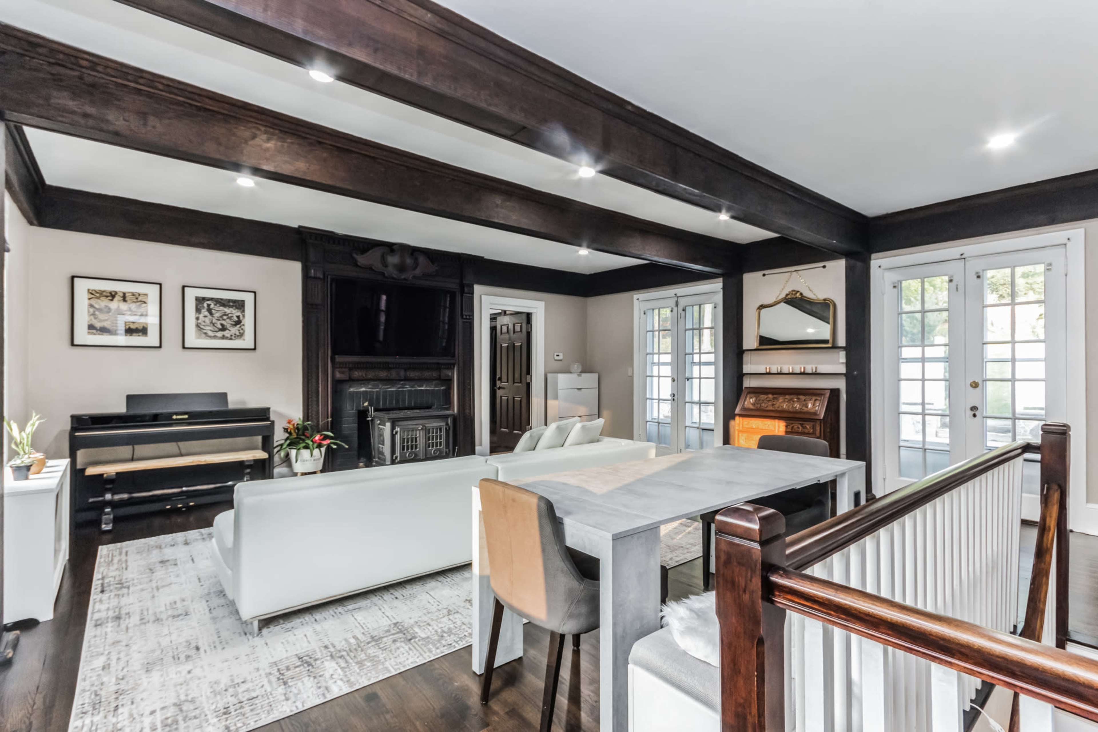 The image shows a spacious living area featuring a white sectional sofa, a wooden dining table, and a piano, with large windows letting in natural light.