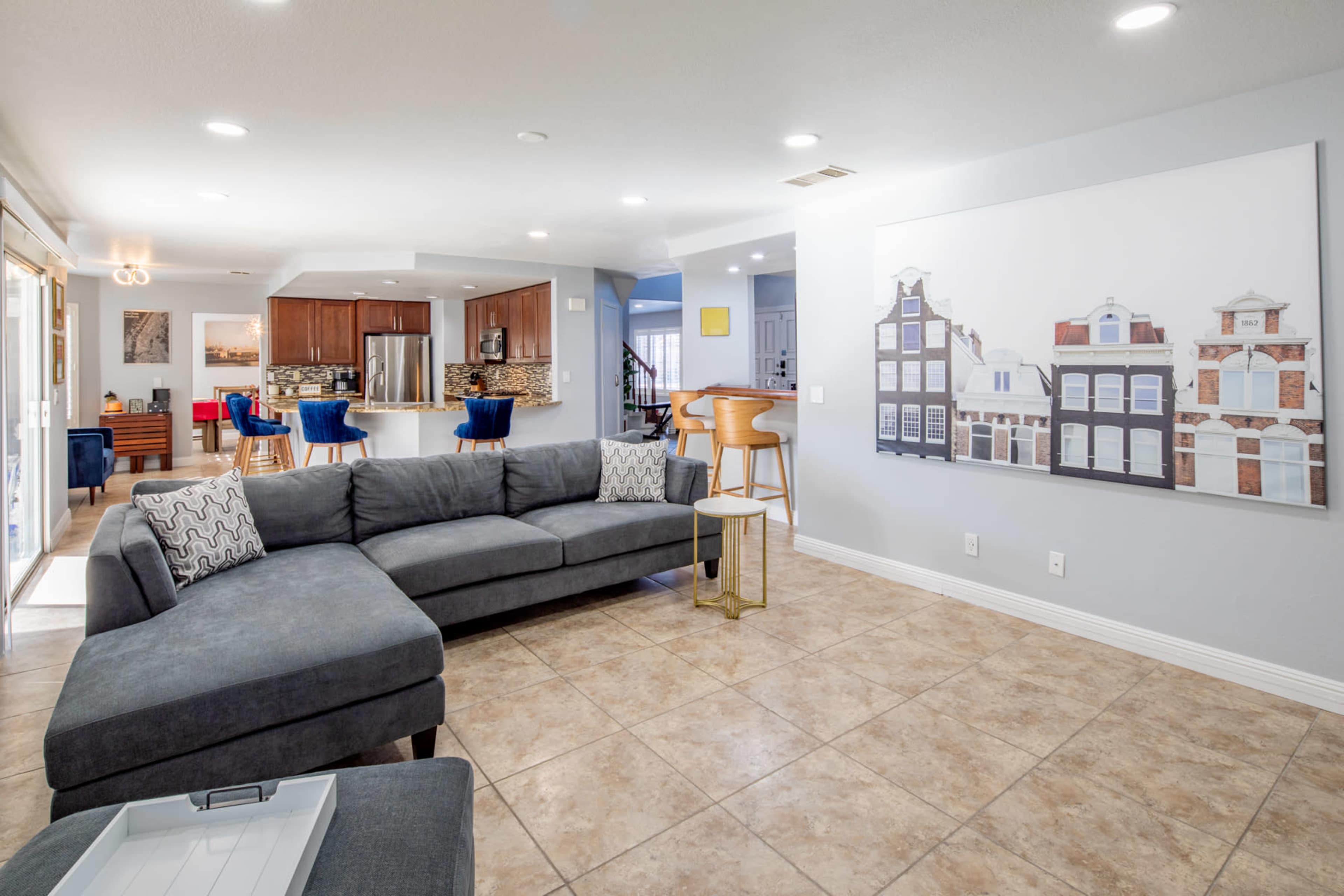 A modern living room features a gray sectional sofa, tiled flooring, and a large artwork on the wall, with an open view to a dining area and kitchen.