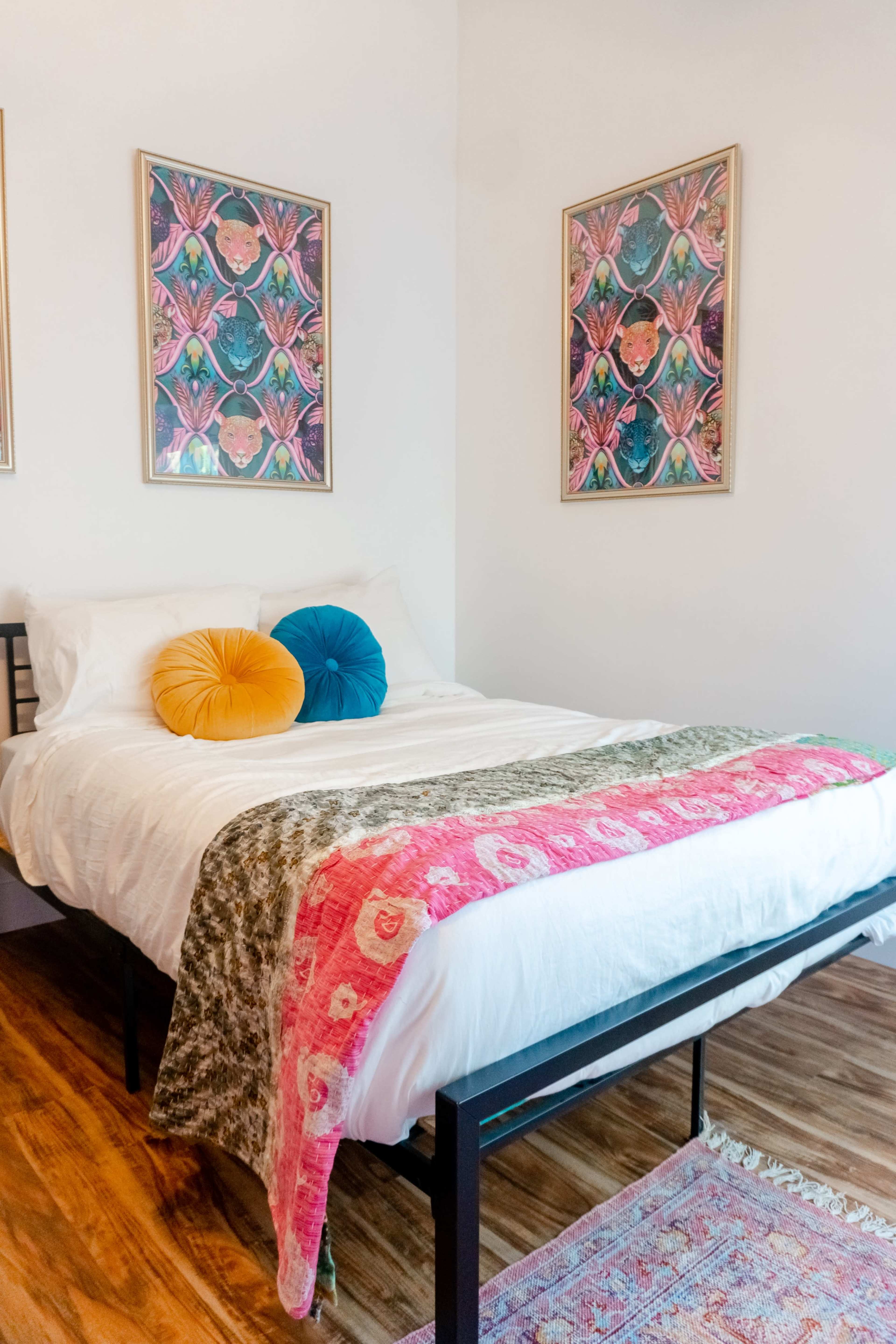 A neatly made bed with colorful decorative pillows and a patterned throw blanket, placed in a well-lit bedroom with two framed artworks on the wall.