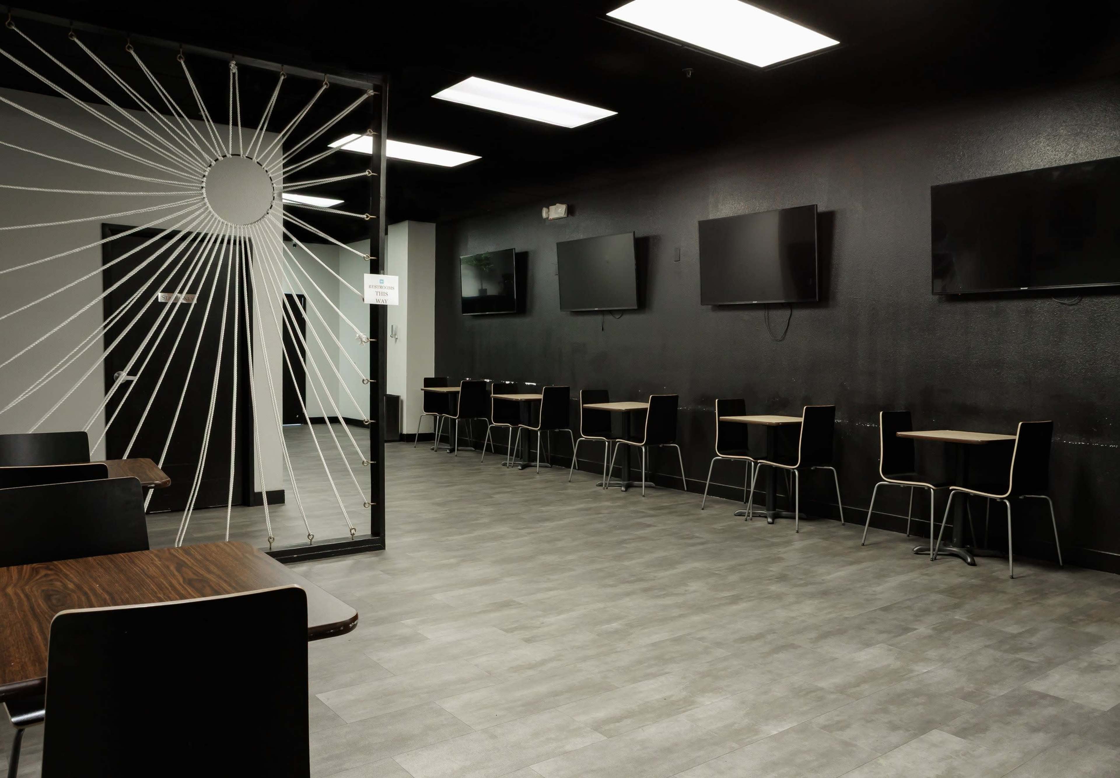 The image shows a modern, empty room with black walls, several flat-screen televisions mounted on one wall, and tables with chairs arranged throughout the space.