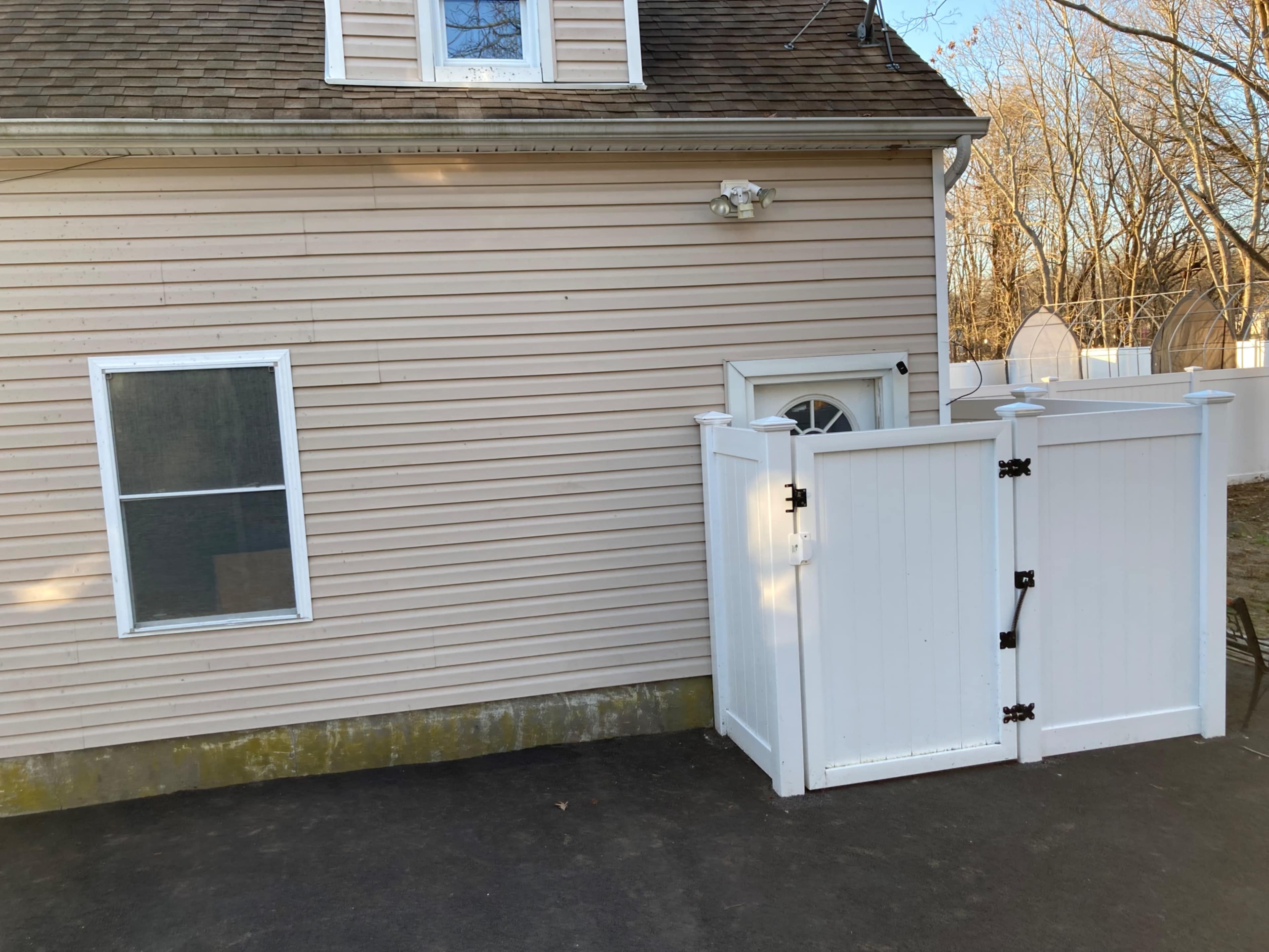 The image shows a beige house with a white gate on the side, leading to a backyard area.