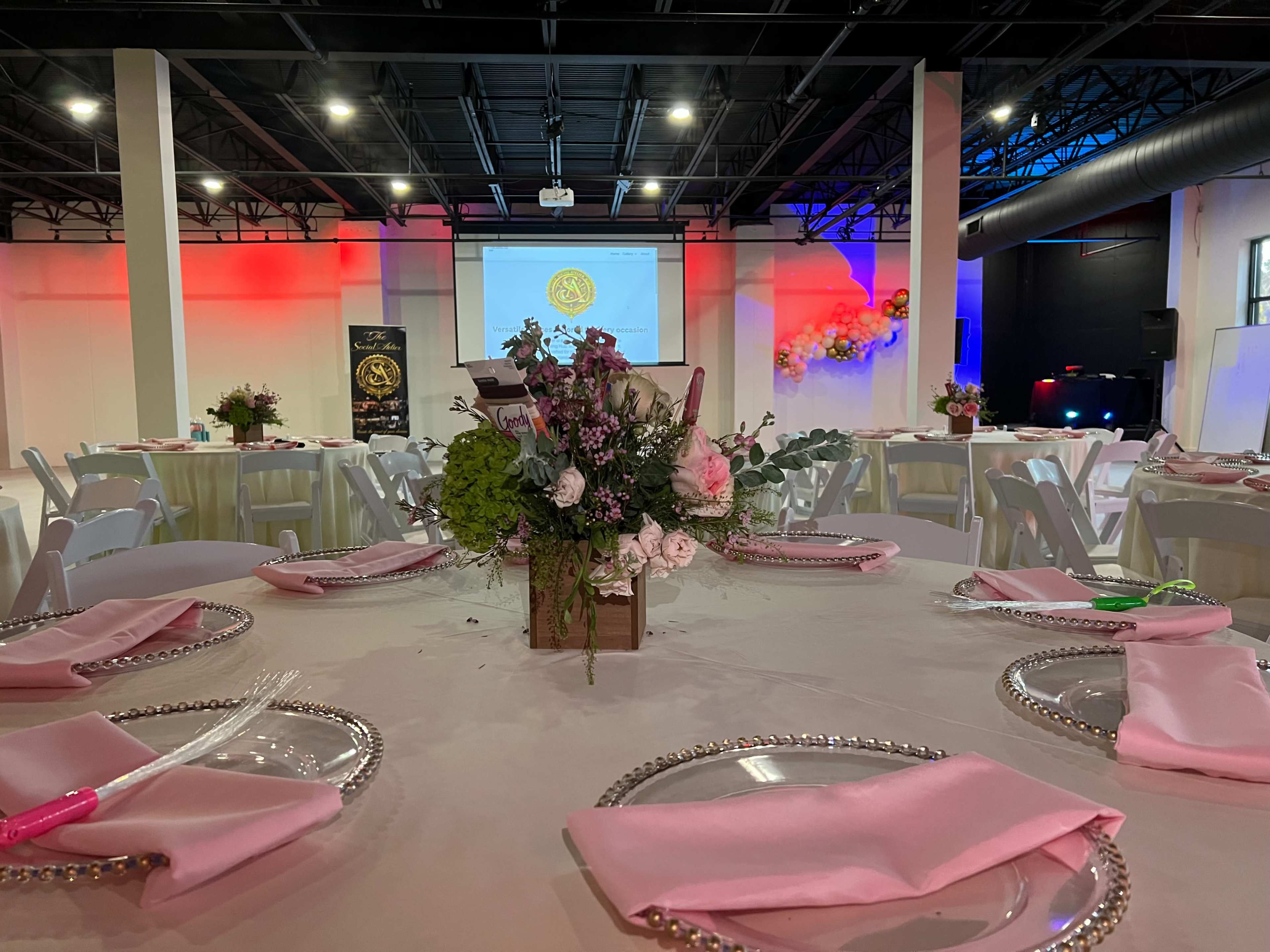 The image shows a banquet hall set up for an event, featuring round tables adorned with floral centerpieces and pink napkins, with a projector screen in the background.