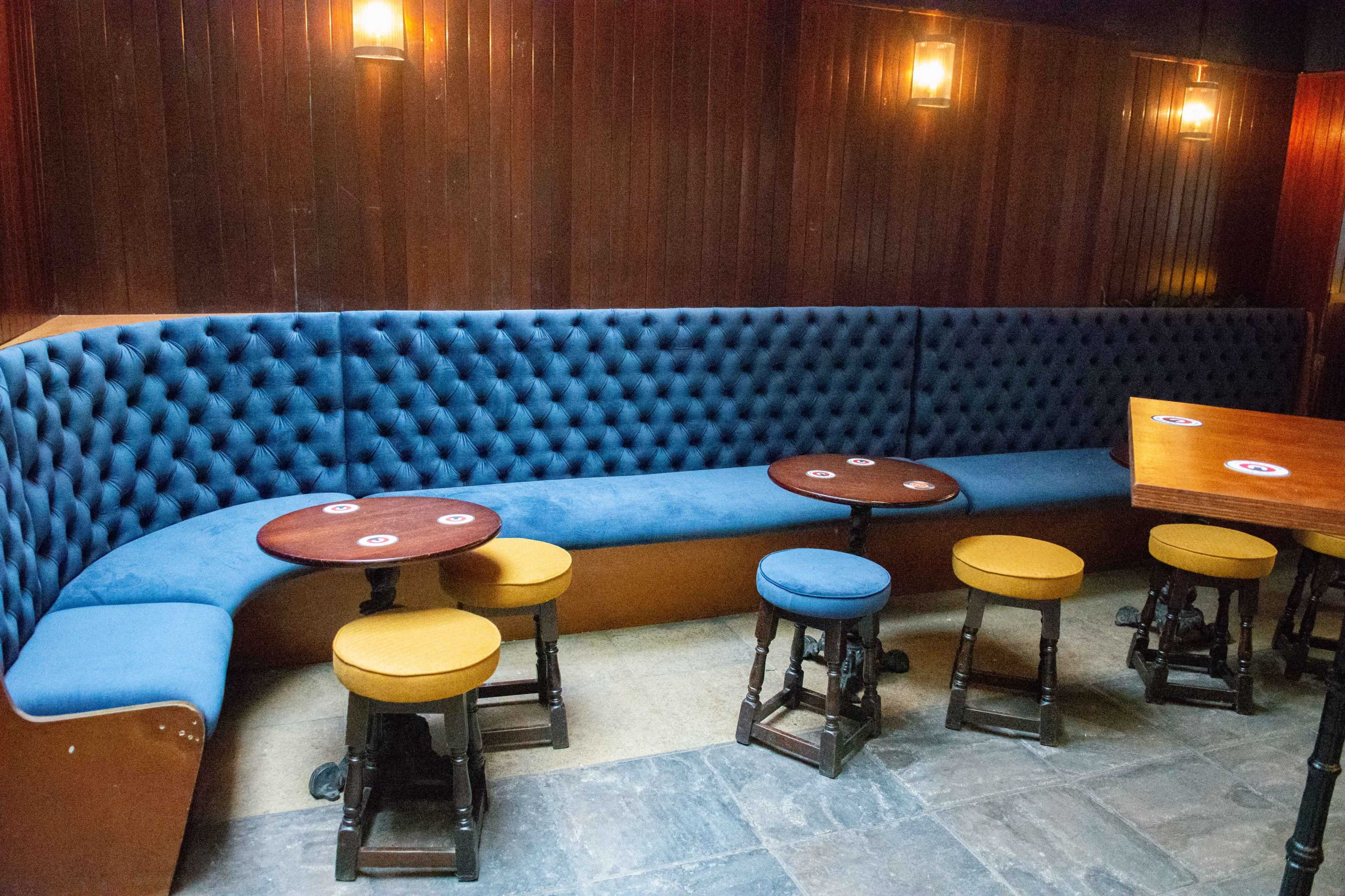 The image shows a cozy interior of a pub with a blue tufted sofa, wooden tables, and round stools with yellow seats.