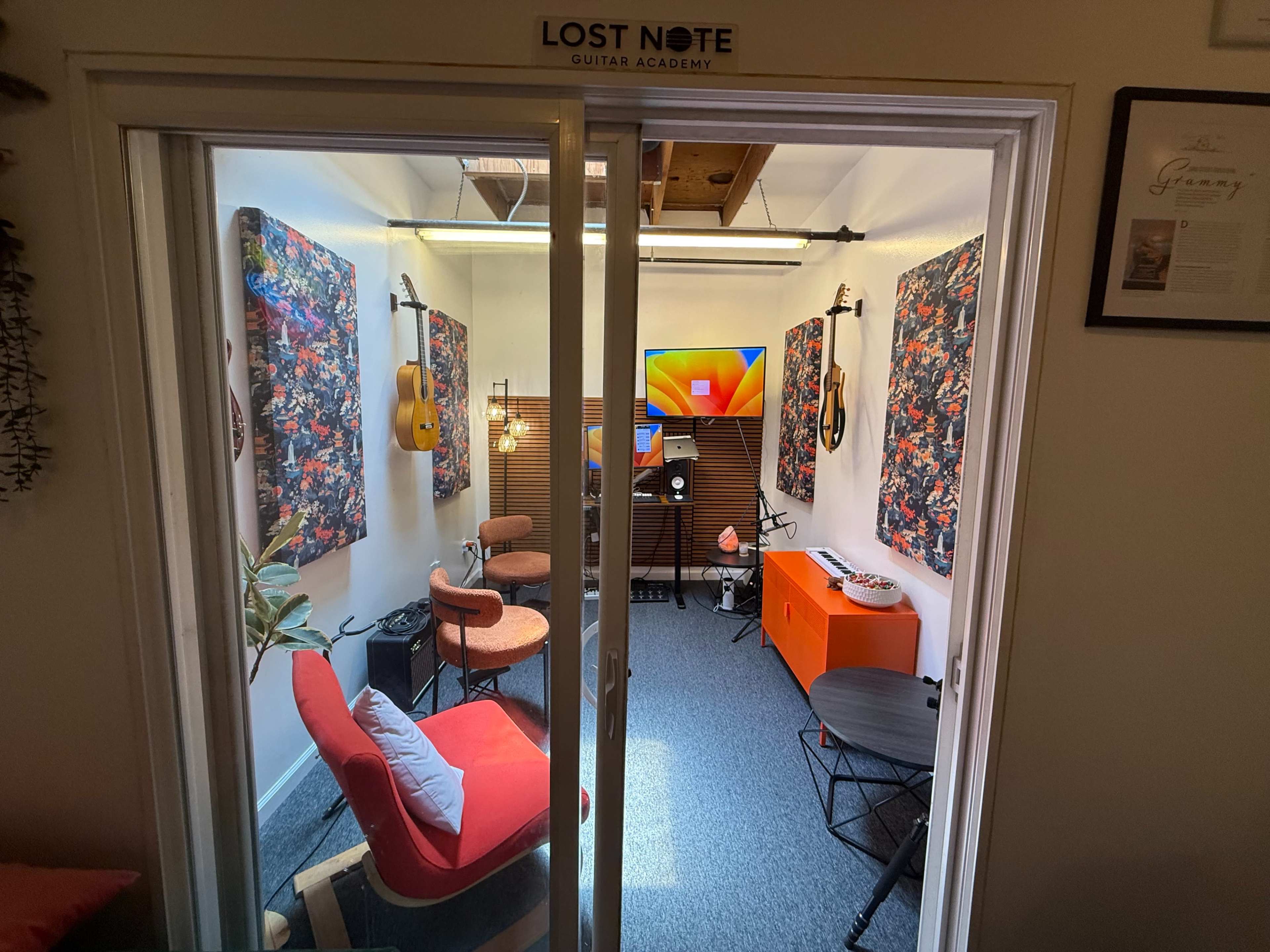 The image shows a small music studio with colorful artwork, musical instruments on the walls, and furniture arranged for sessions, labeled as "Lost Note Guitar Academy."