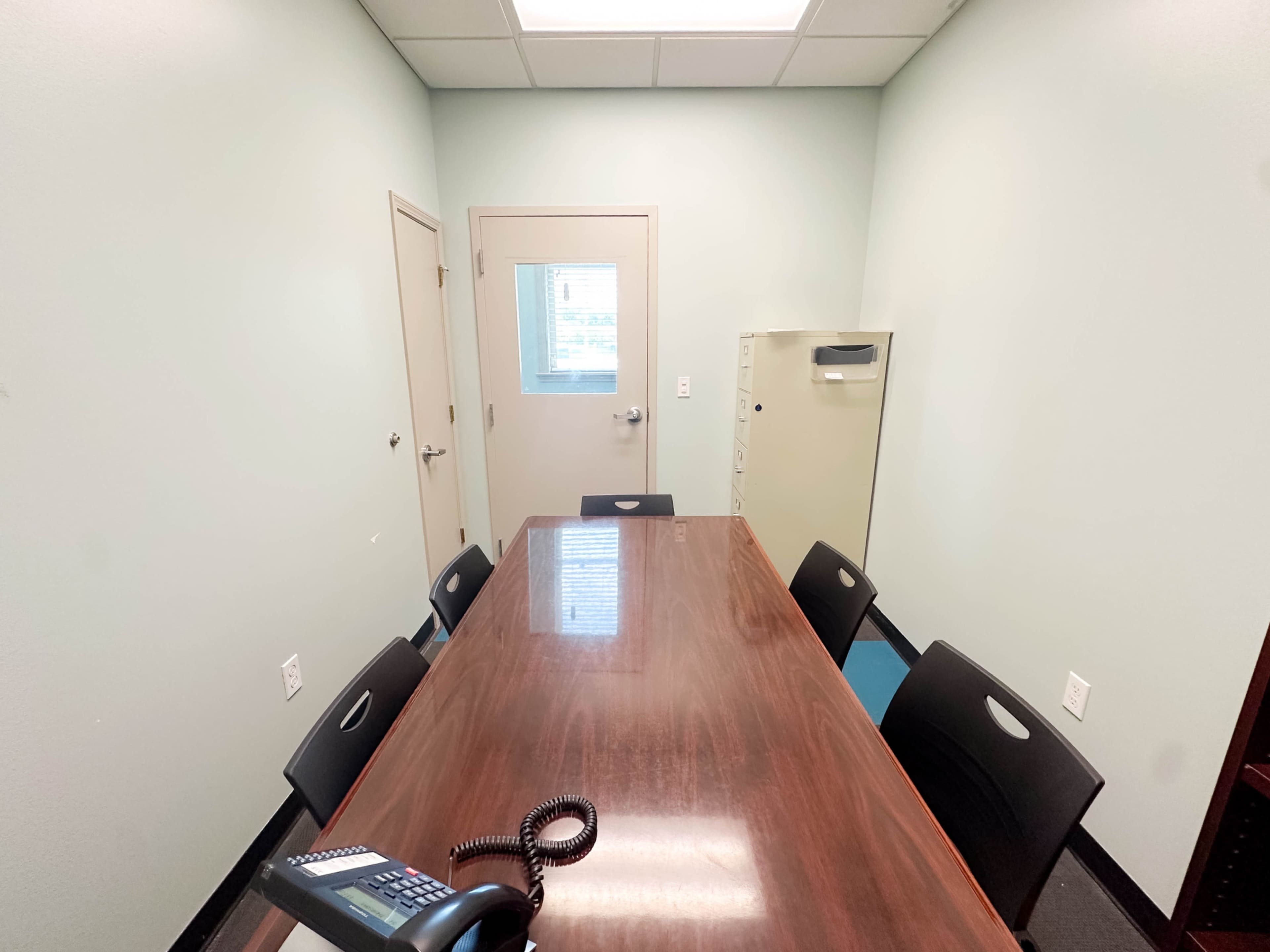 The image shows a small, rectangular meeting room with a wooden table surrounded by six black chairs, a phone on the table, and a file cabinet in the corner.