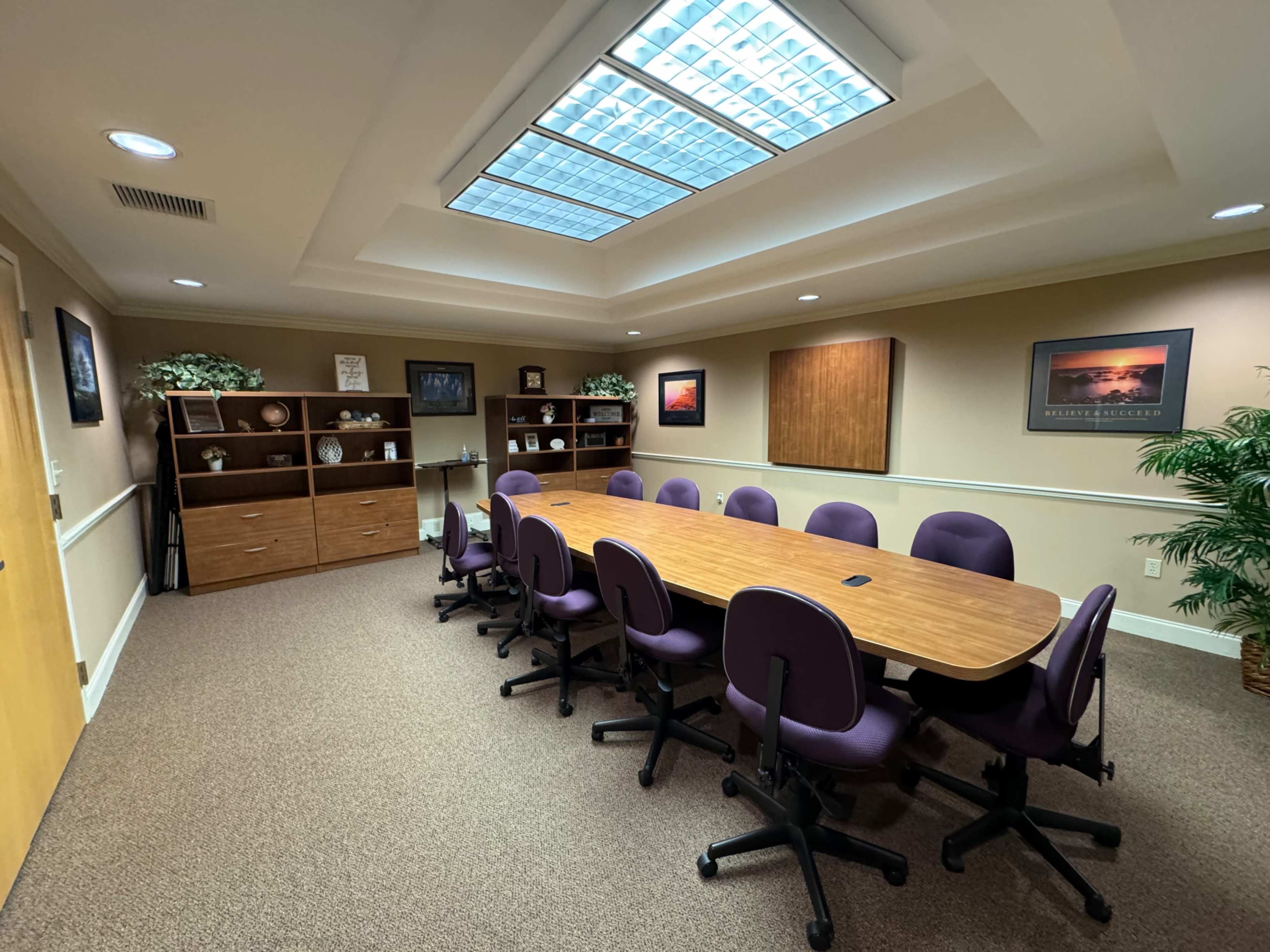 A conference room features a long wooden table surrounded by purple office chairs, with storage cabinets and a skylight above.