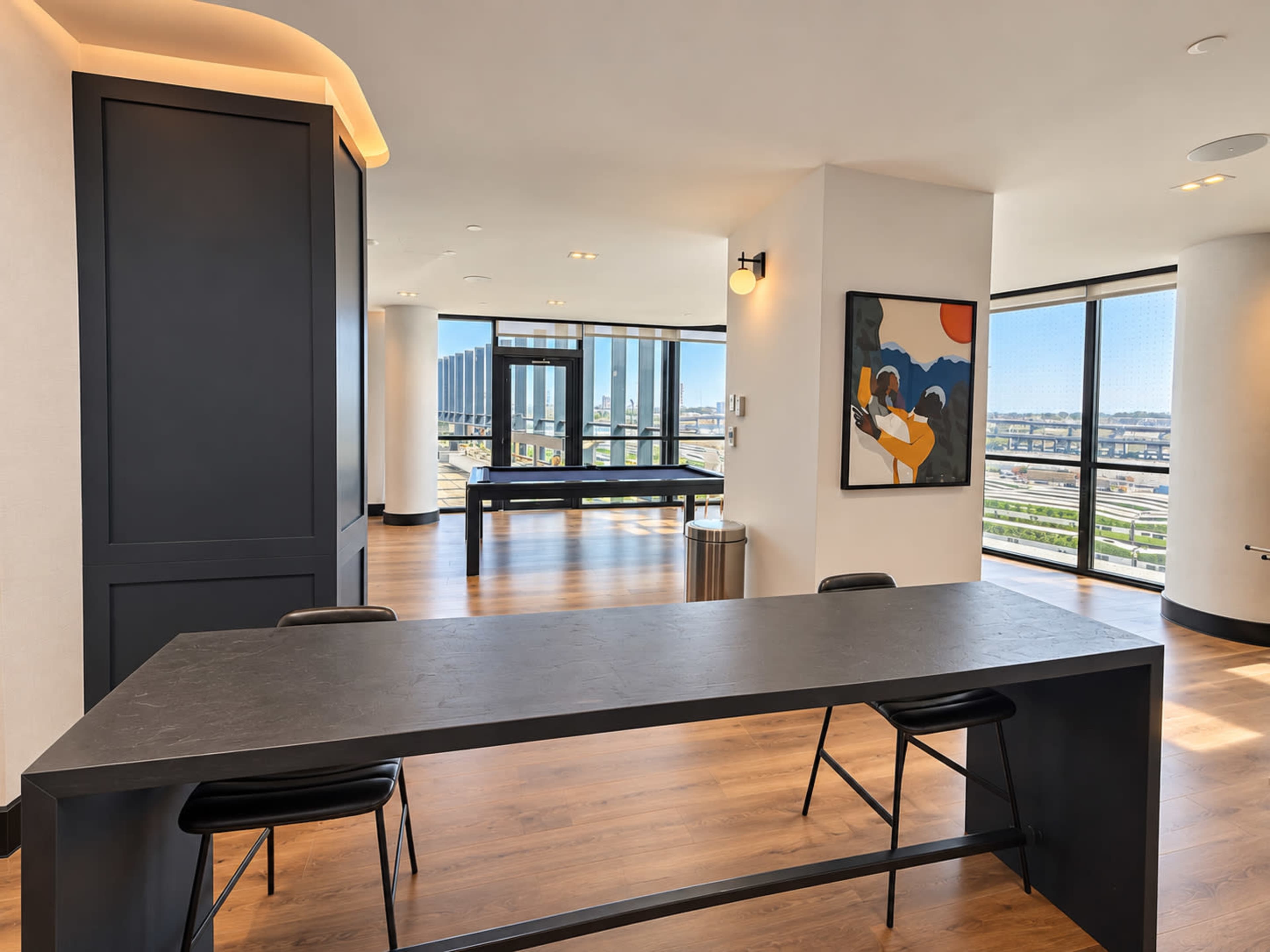 The image shows a modern interior with large windows, featuring a long black table, black chairs, and an abstract painting on the wall.