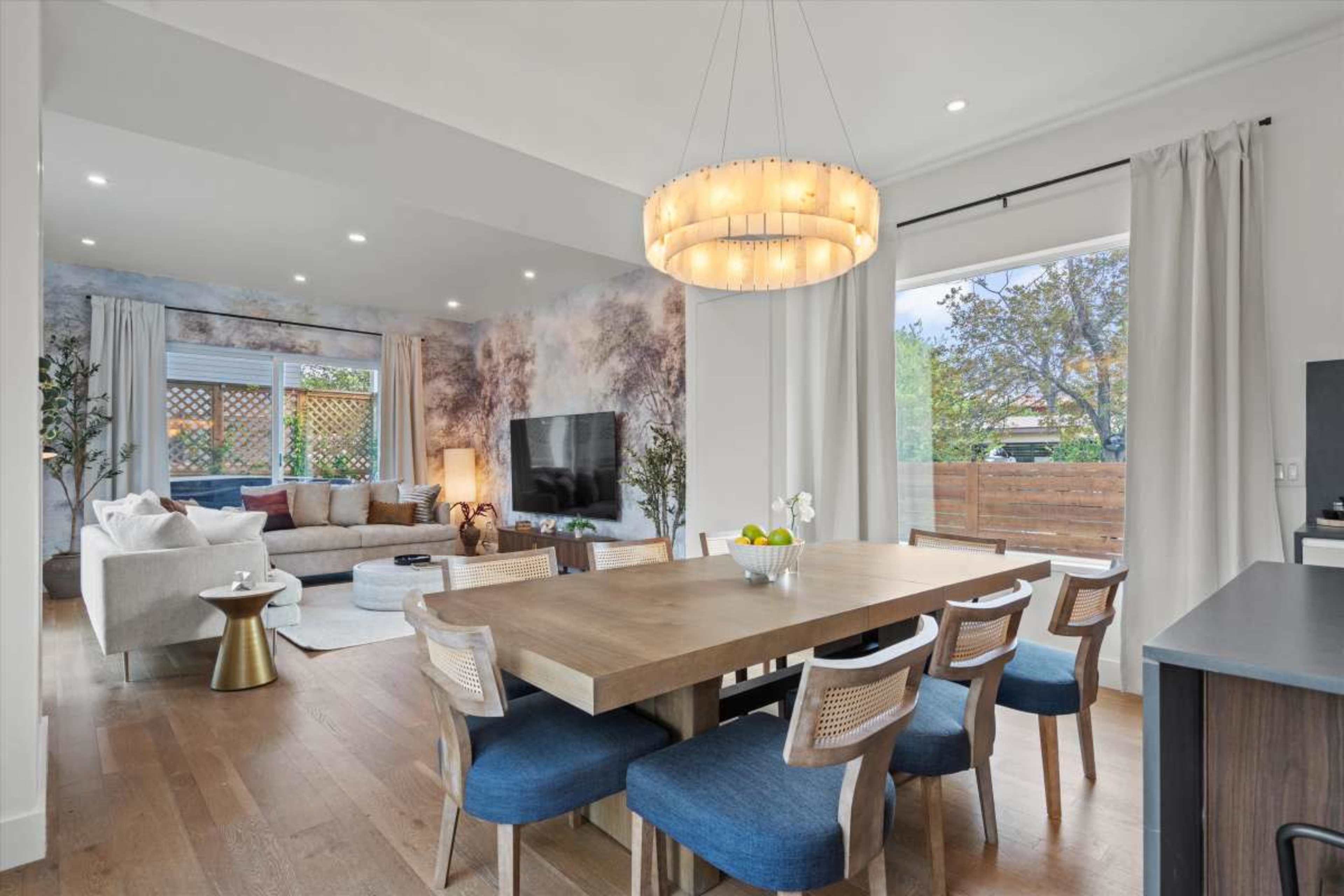 A dining area with a large wooden table and blue chairs is adjacent to a contemporary living space featuring a sofa, television, and large windows.