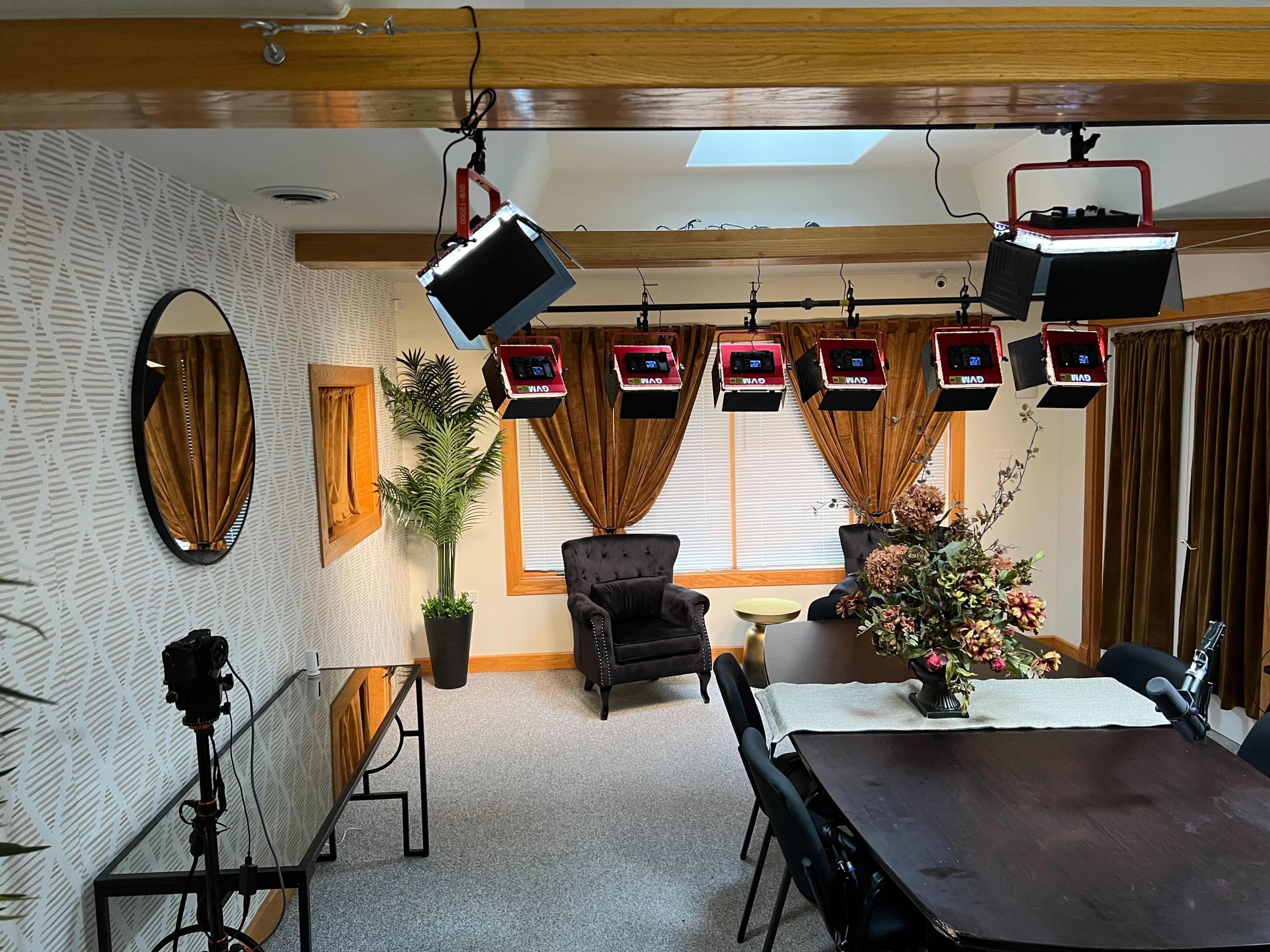 A well-lit room featuring a dining table, a black armchair, and several hanging studio lights above.