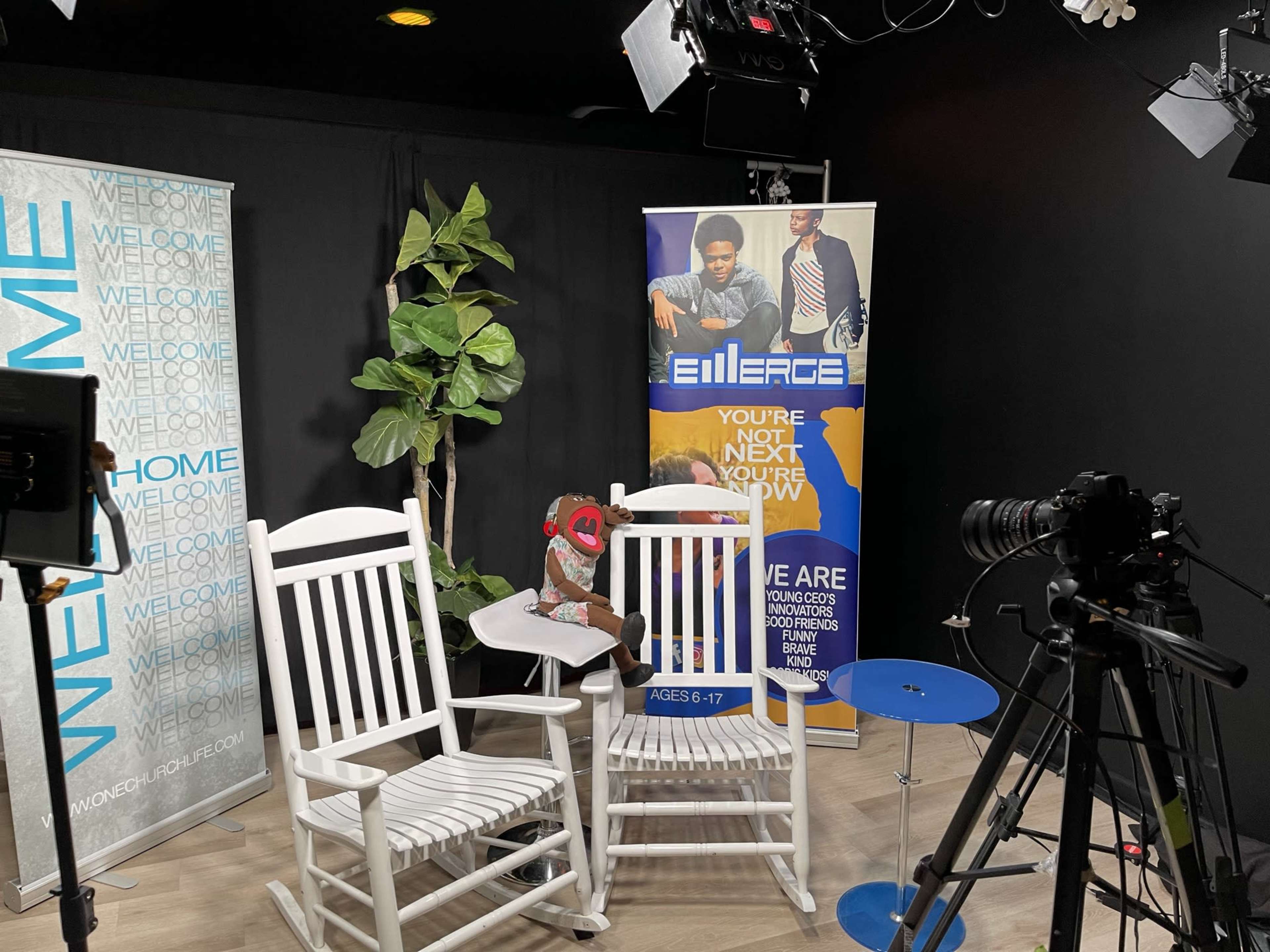 A small studio setup with two white rocking chairs, a blue side table, a plant, and signage promoting a youth program.
