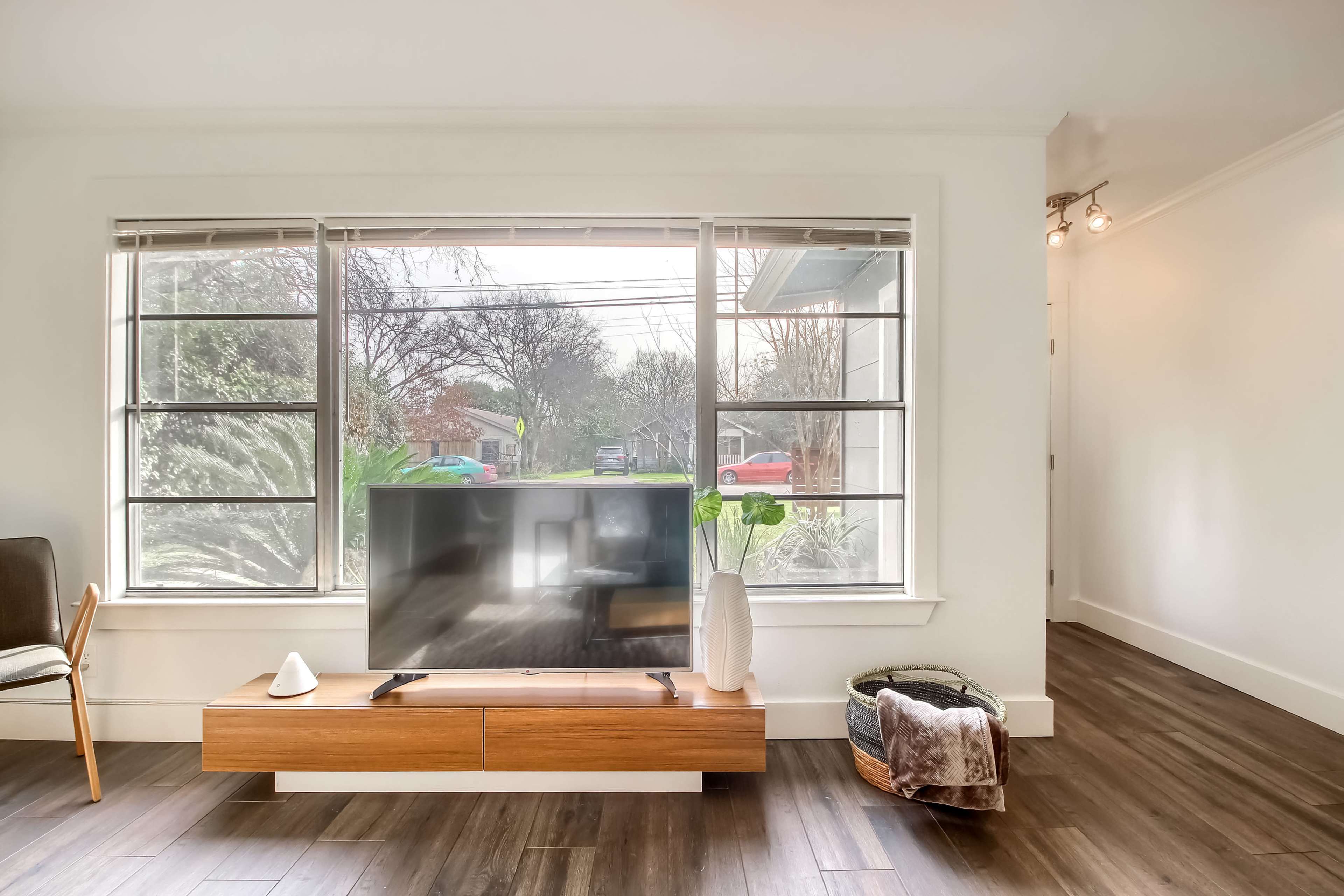 A living room features a television on a wooden media console, complemented by large windows allowing natural light to enter.