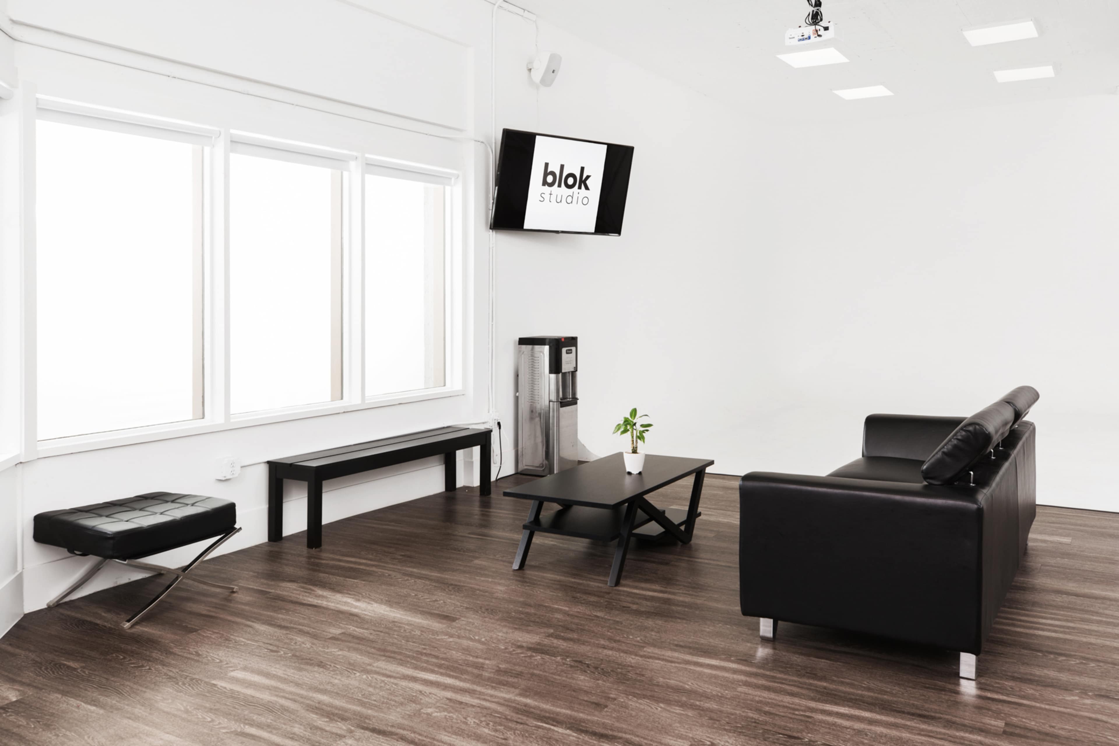 The scene depicts a minimalist studio interior with a black leather sofa, a coffee table, a bench, and large windows, featuring a water dispenser and a mounted screen displaying "blok studio."