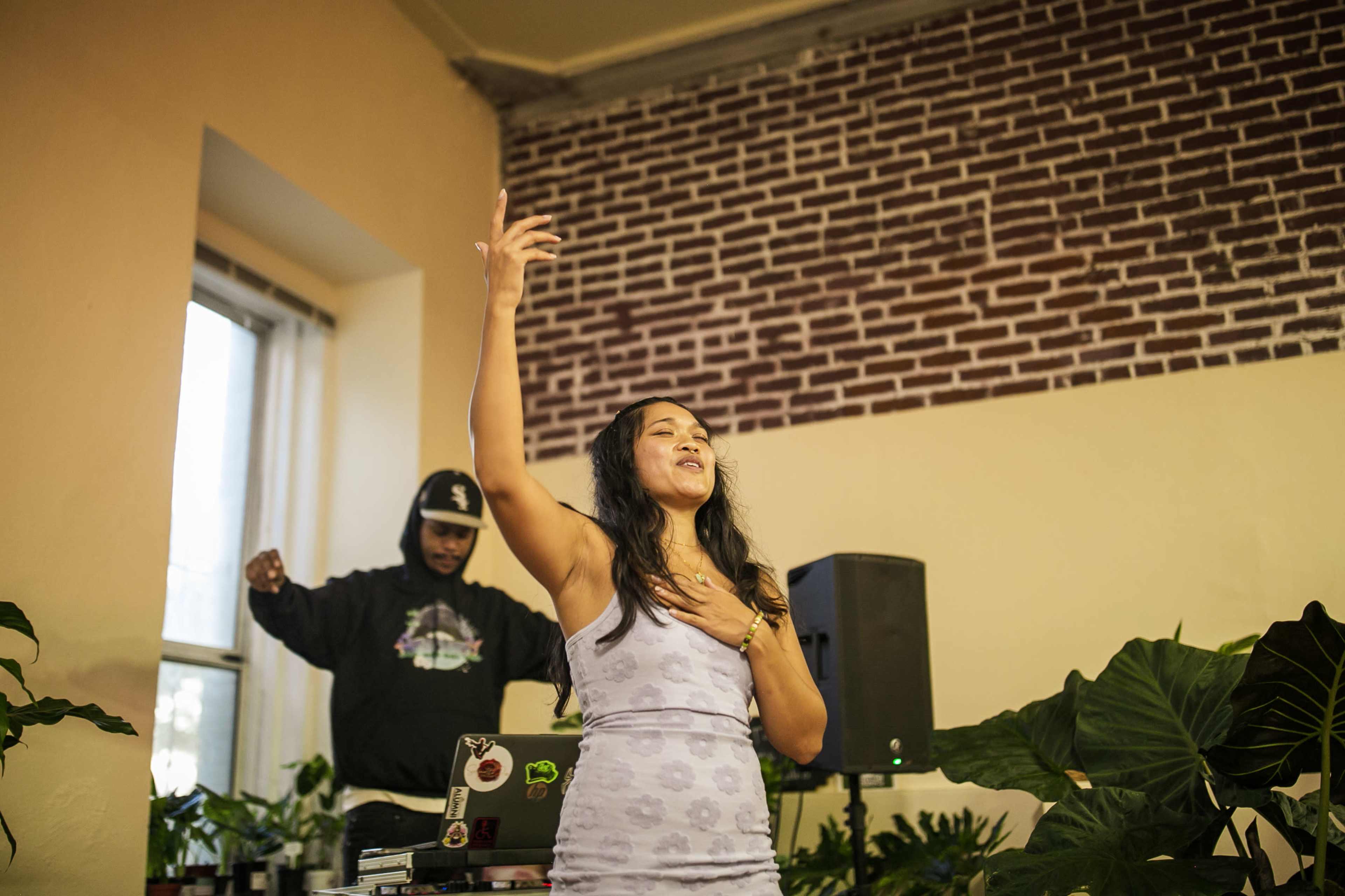 A woman sings on stage with a DJ in the background, surrounded by plants in a warmly lit room.