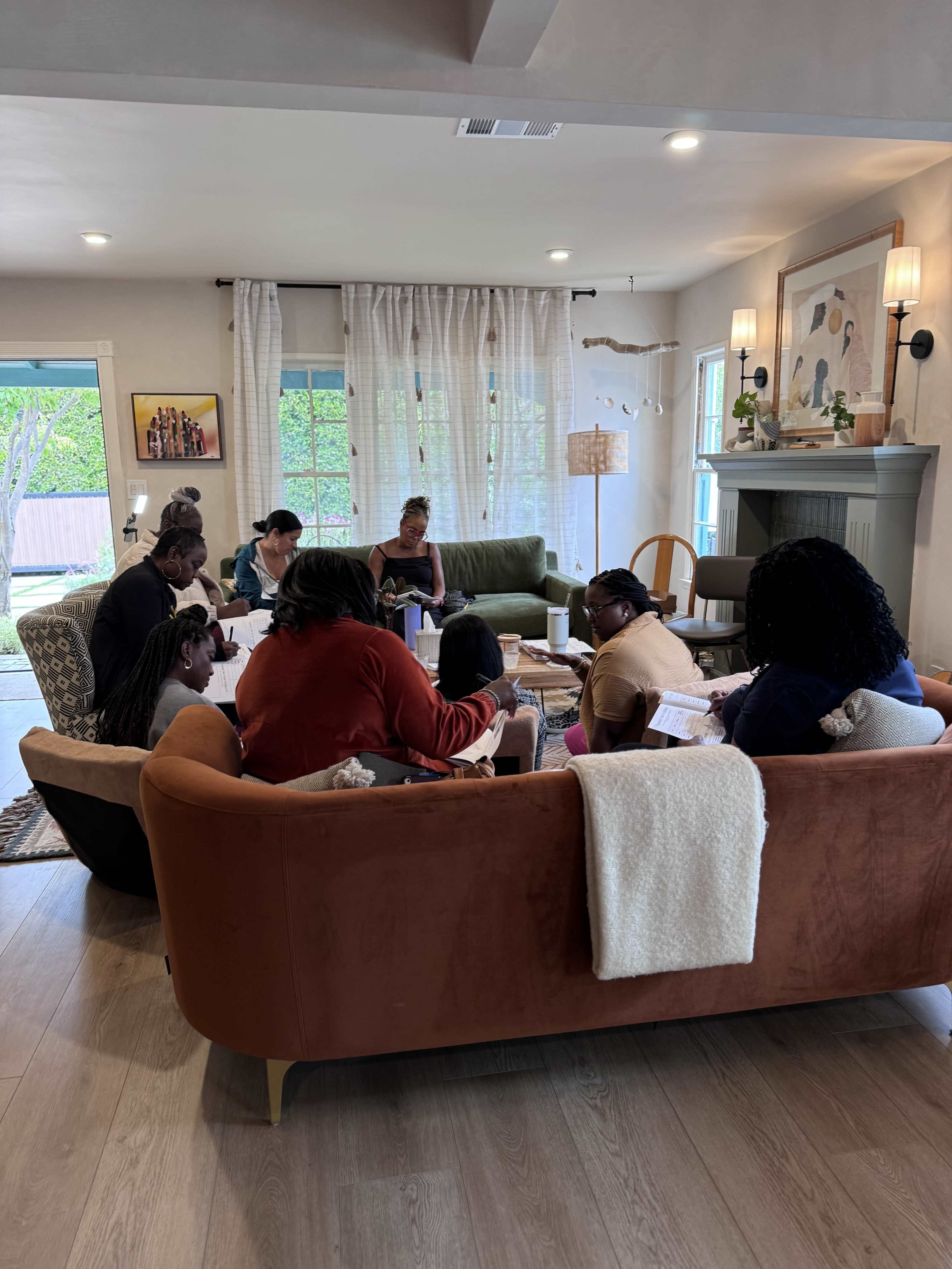 A group of women is gathered in a living room, engaged in conversation while seated on a large, orange couch.