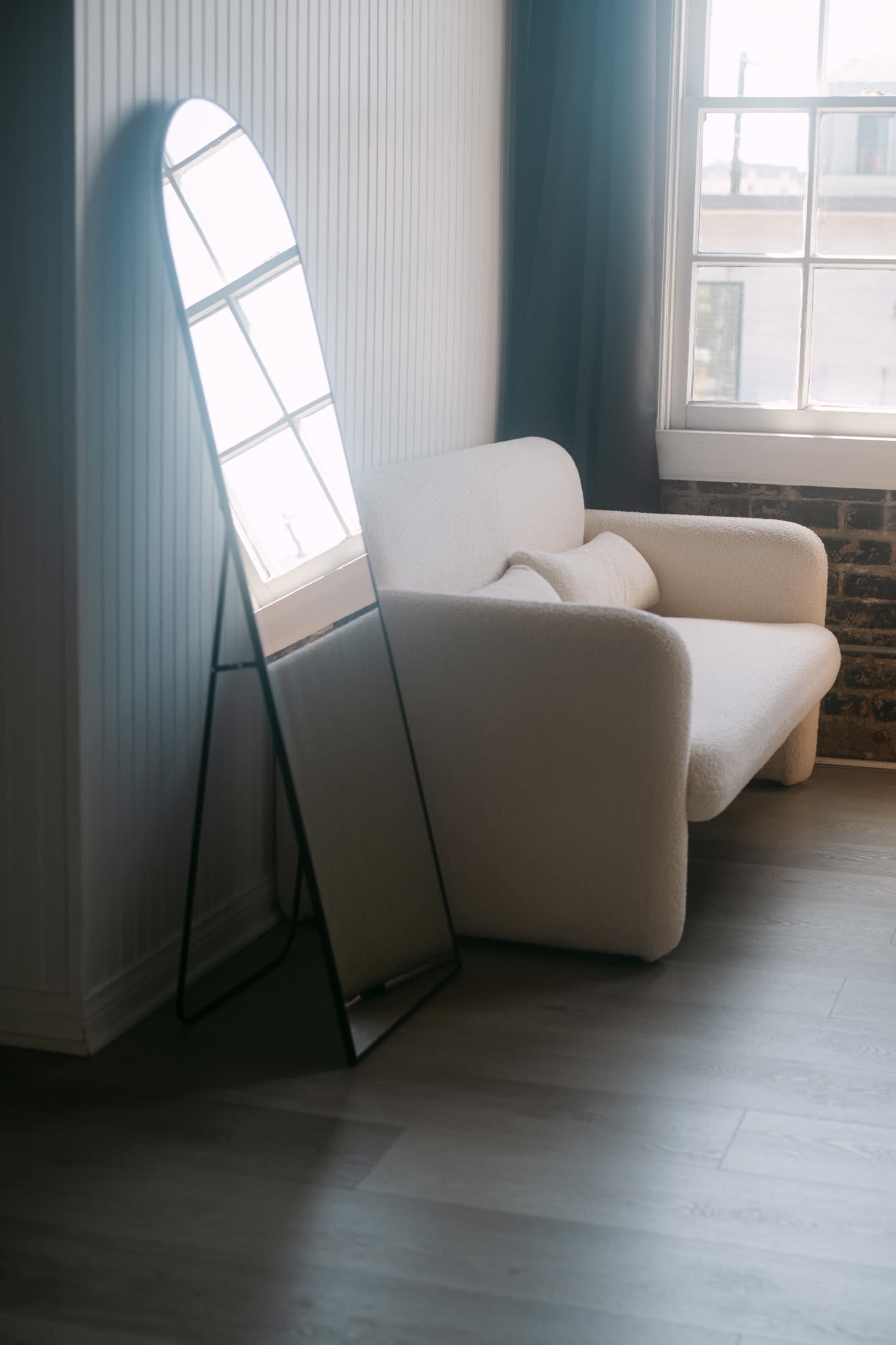 A cozy corner features a plush armchair next to a tall, standing mirror with a sleek frame, illuminated by natural light from a nearby window.