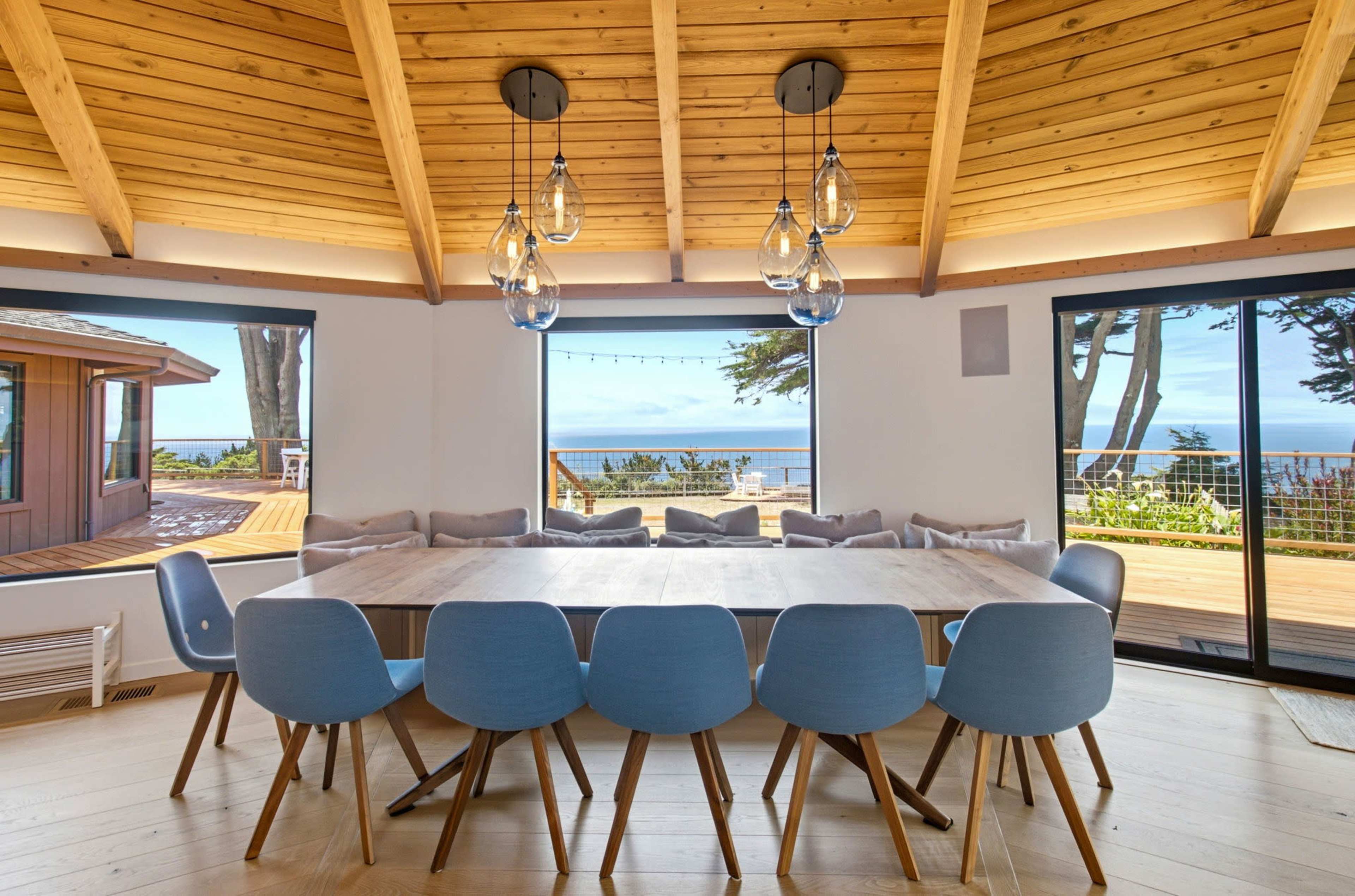 A spacious dining area features a large wooden table surrounded by blue chairs, with large windows offering a view of the outdoors.