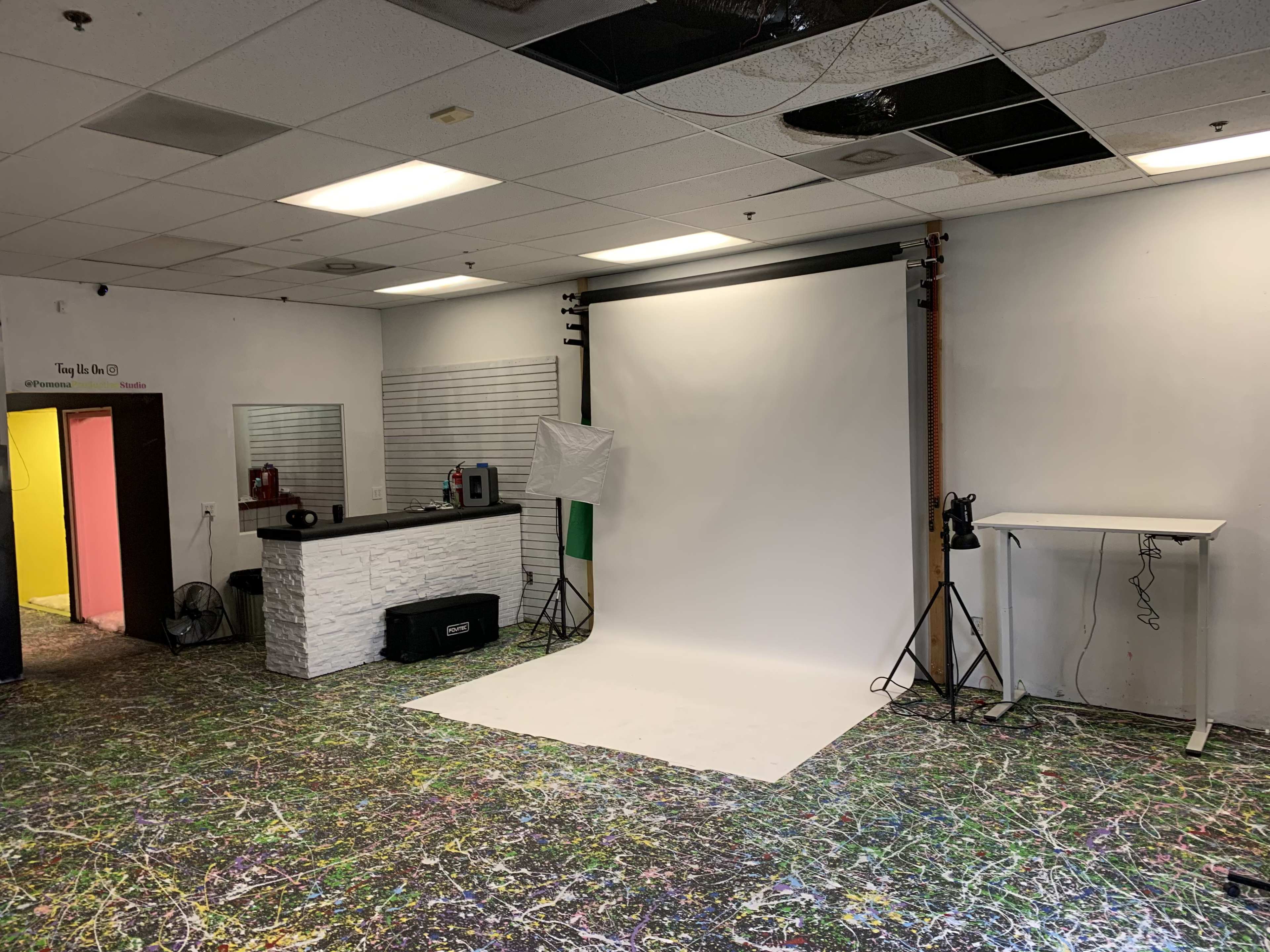 The image shows a photography studio with a white backdrop, colorful splatter-painted floor, and a counter with studio equipment.