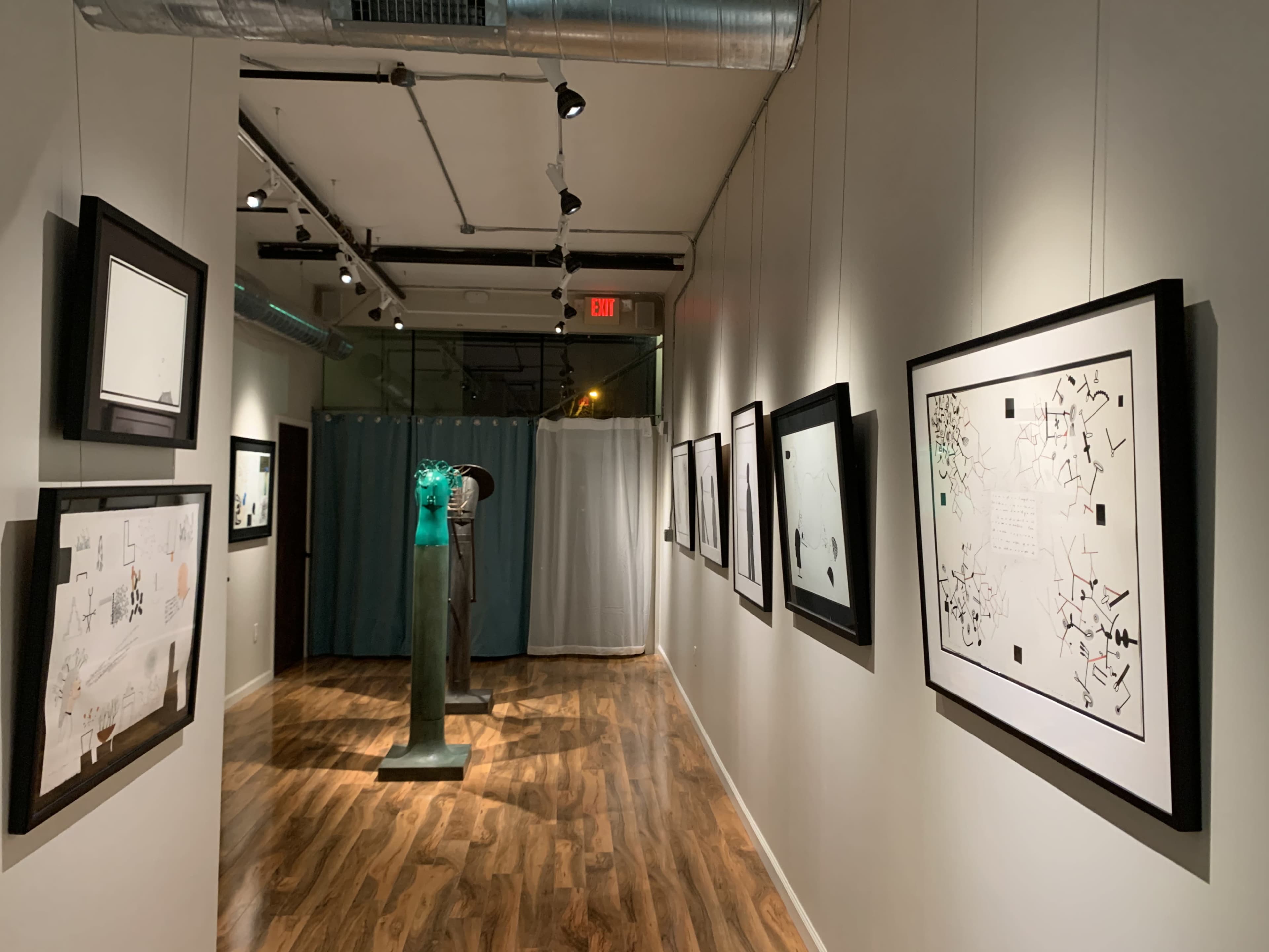 The image shows a gallery hallway with framed artworks on the walls and a sculpture at the far end, partially obscured by a curtain.