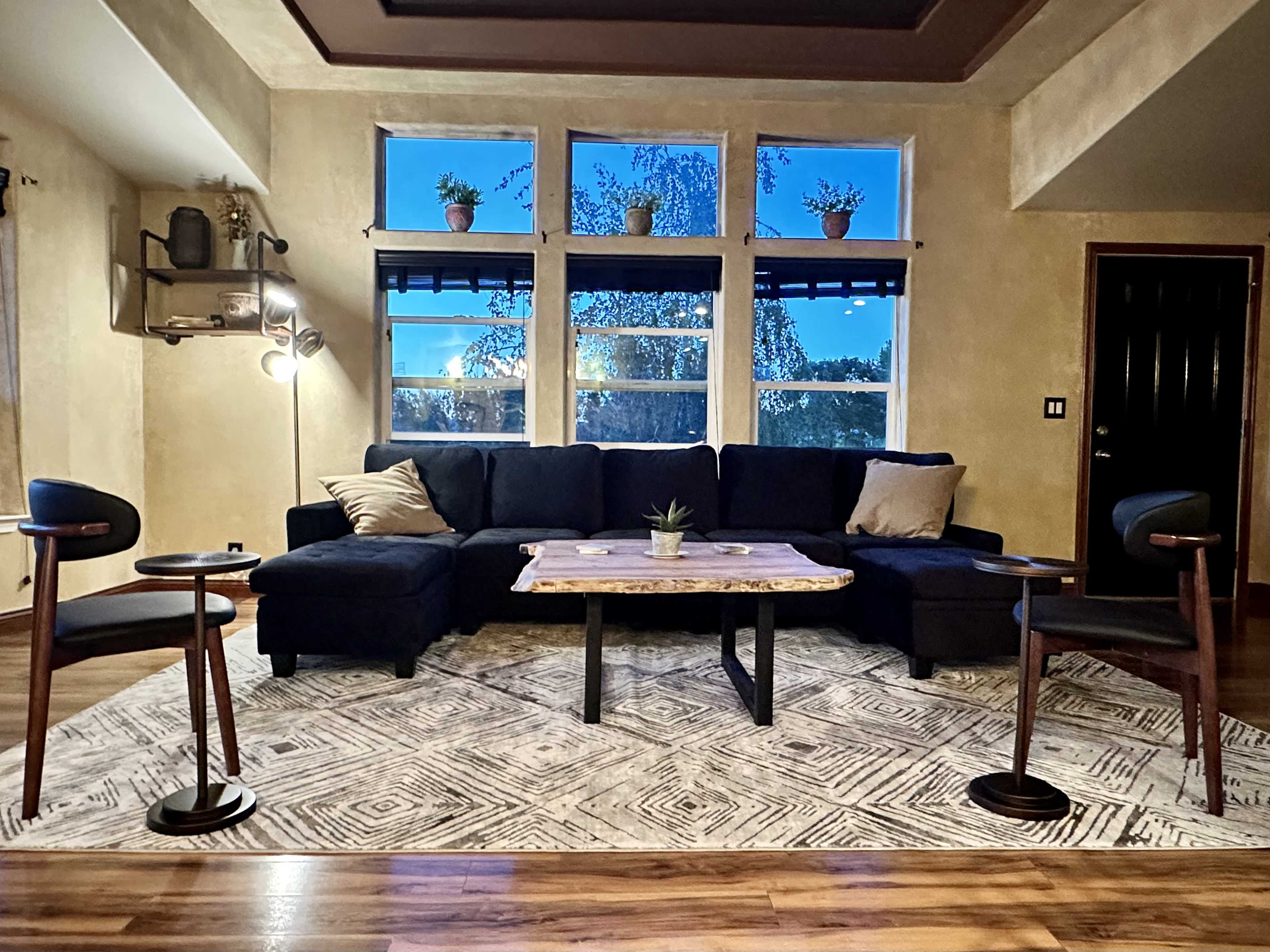 A modern living room features a navy sectional sofa, a wooden coffee table with a succulent, and three high-backed chairs surrounding it, with large windows revealing an evening sky.