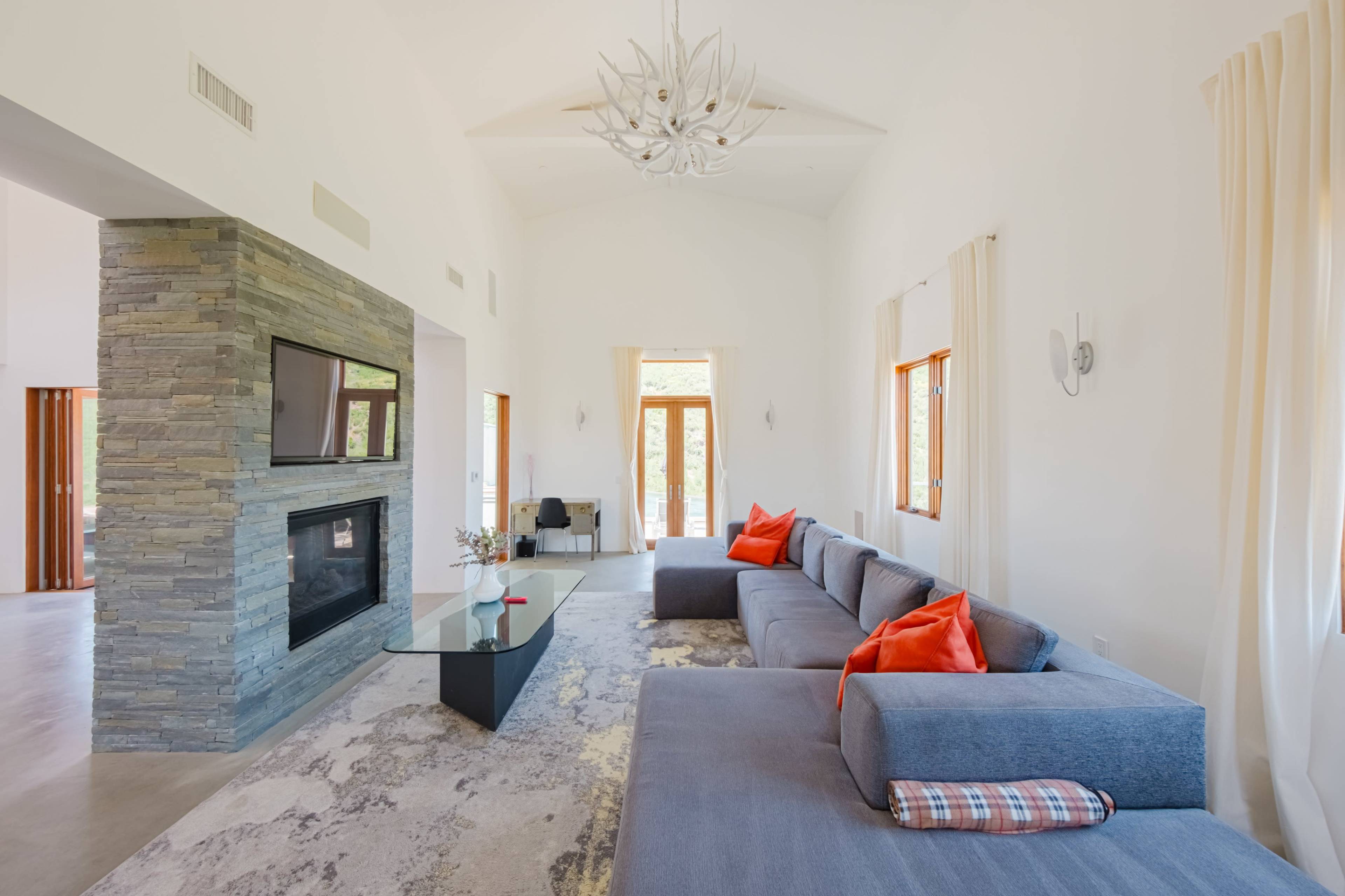 A modern living room with a stone fireplace, a large gray sectional sofa adorned with orange cushions, and large windows providing natural light.