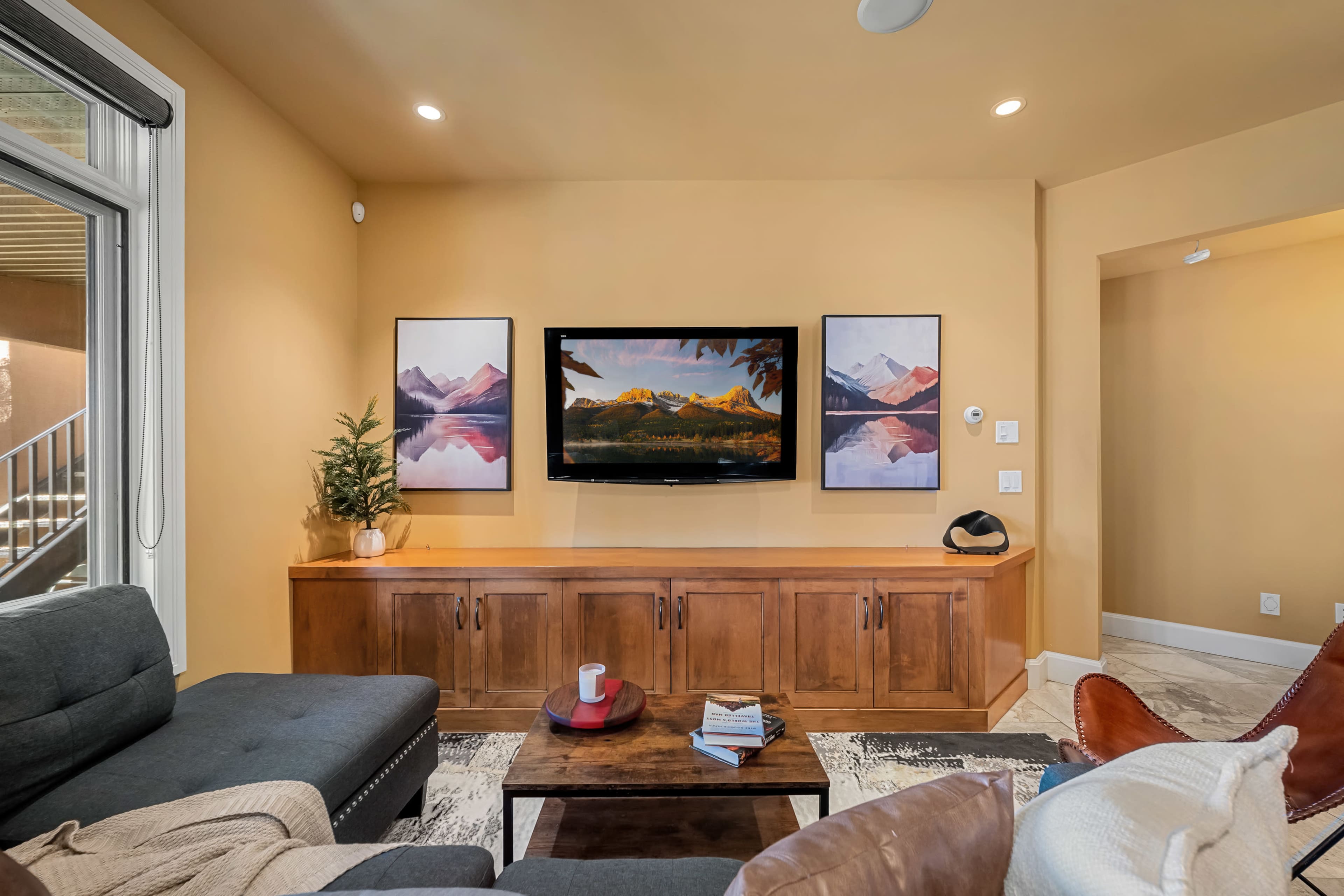 The image shows a living room with a television mounted on a wall above a wooden cabinet, flanked by two framed landscape pictures and furnished with a sofa and a coffee table.
