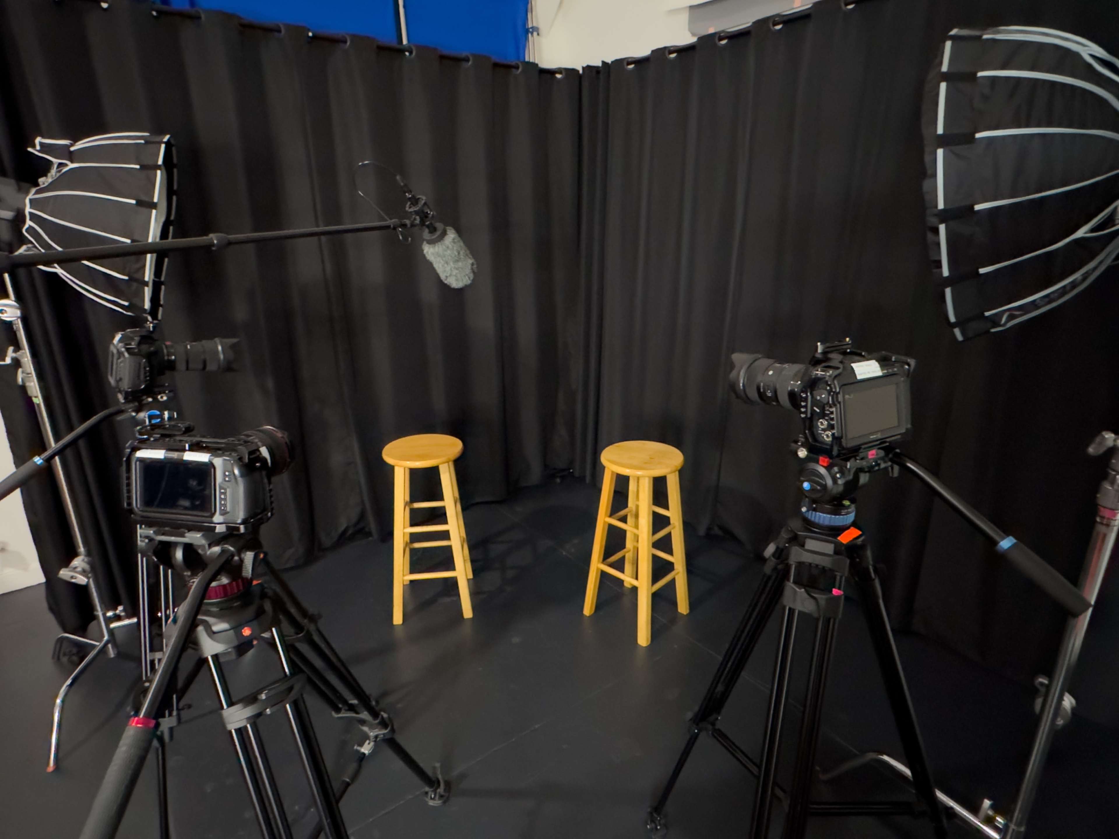 The image shows a small studio setup featuring two wooden stools, flanked by cameras and microphones, with black curtains in the background.