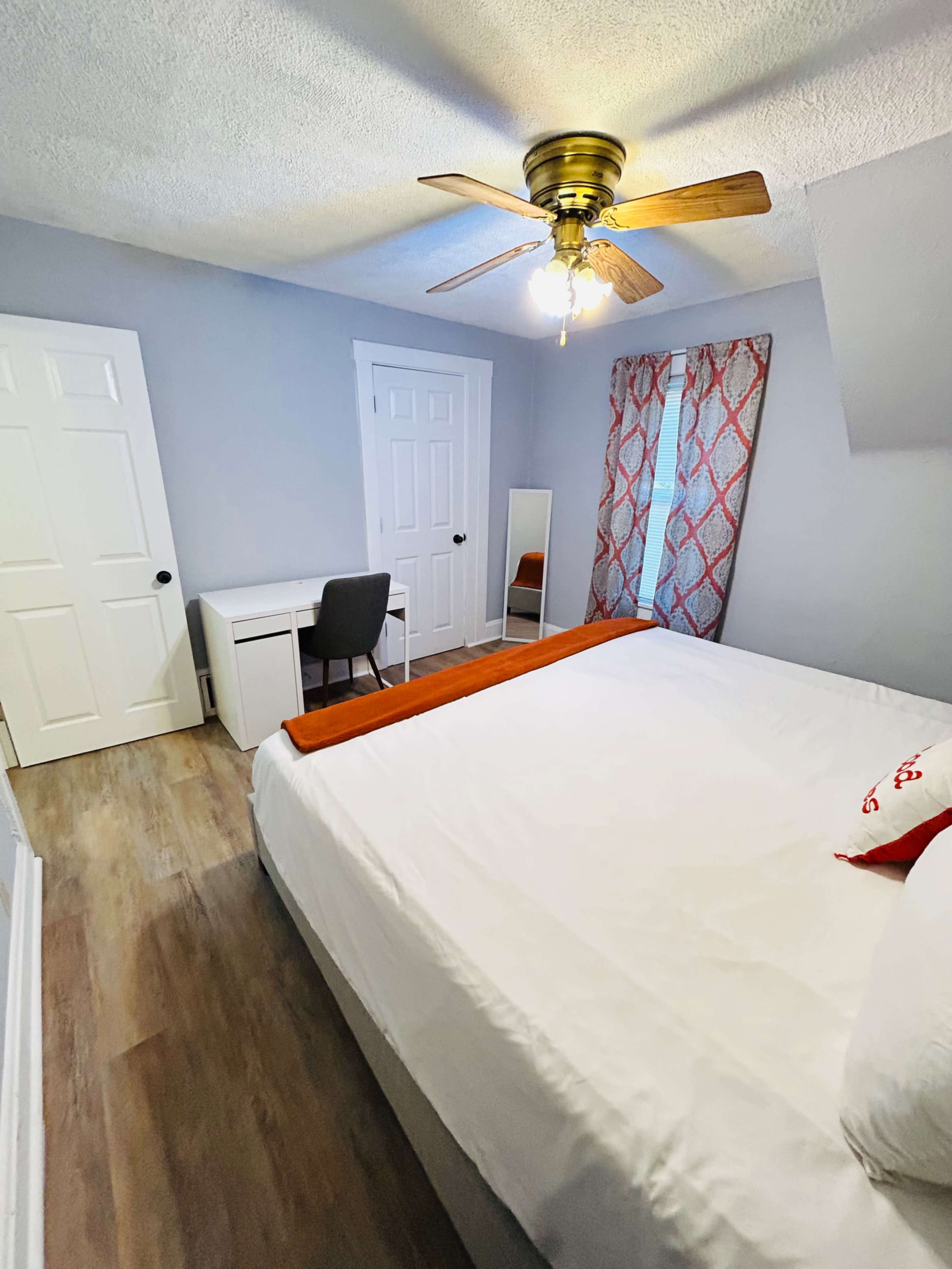 A small bedroom featuring a bed, a desk with a chair, a mirror, a window with patterned curtains, and doors leading to a closet and the outside.