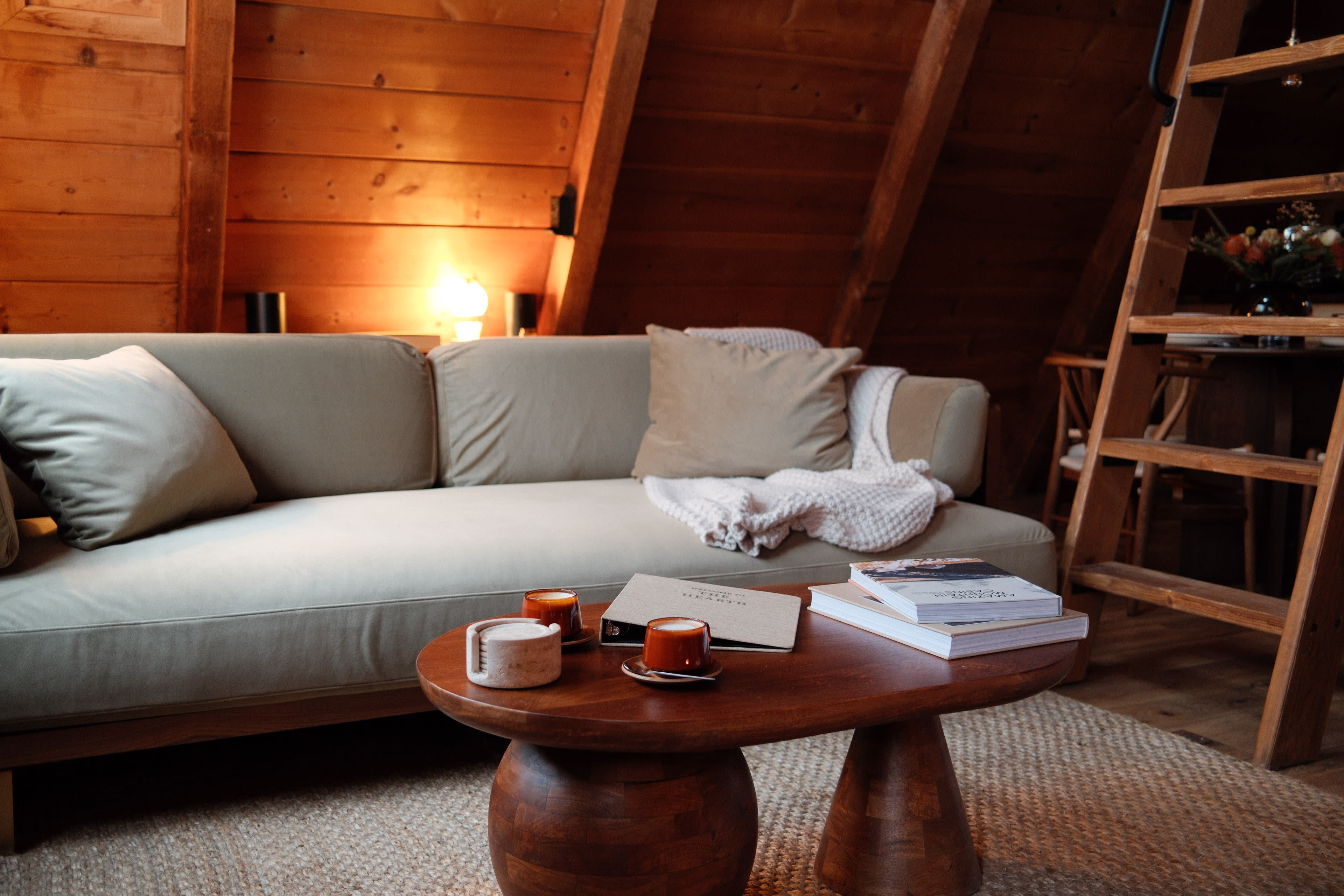 A cozy wooden attic living space features a beige couch, a wooden coffee table with two orange cups, and a stack of books.