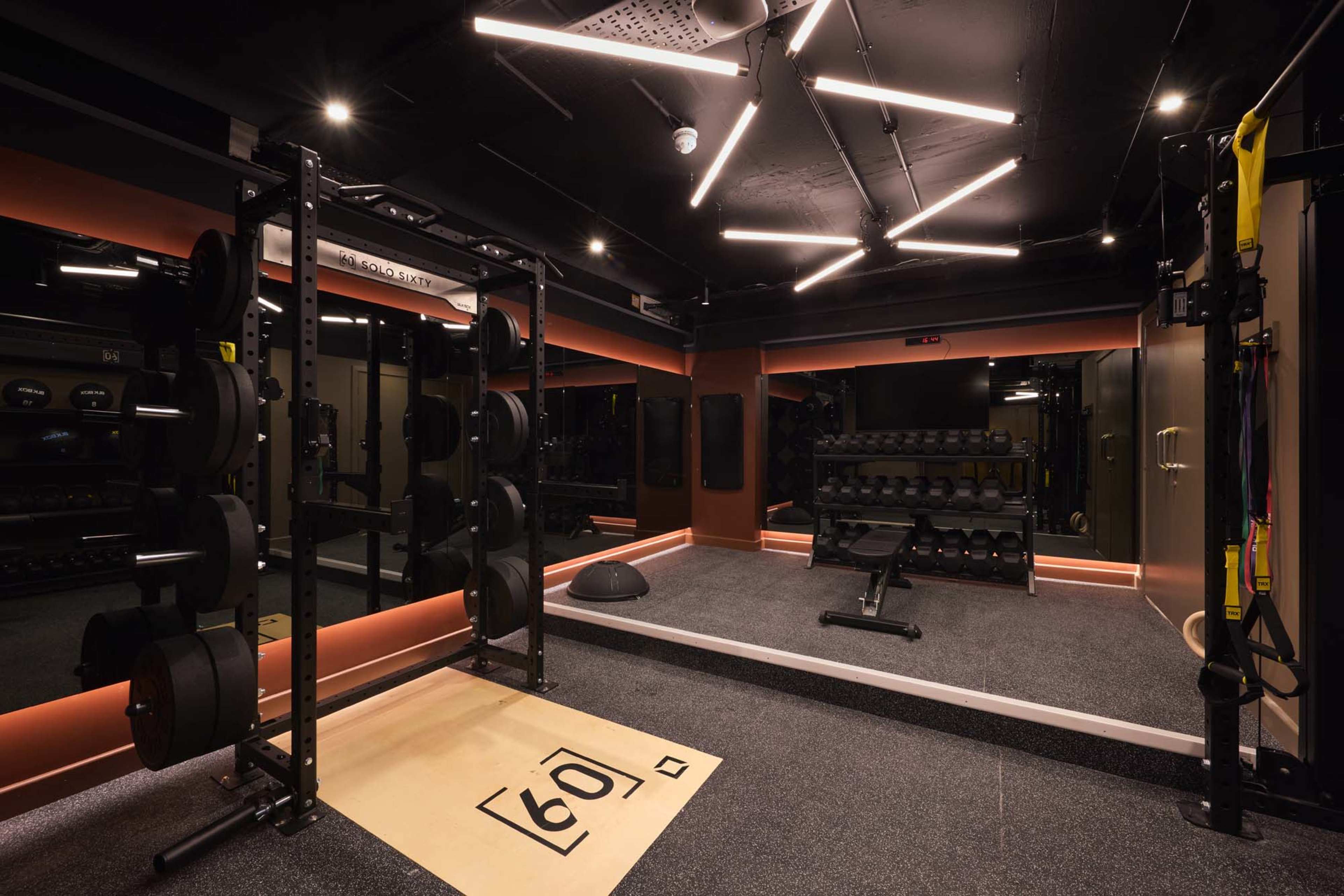 The image shows a modern gym interior equipped with weightlifting equipment, including a squat rack and dumbbell racks, along with a functional training area featuring a rubber floor and ceiling lights.