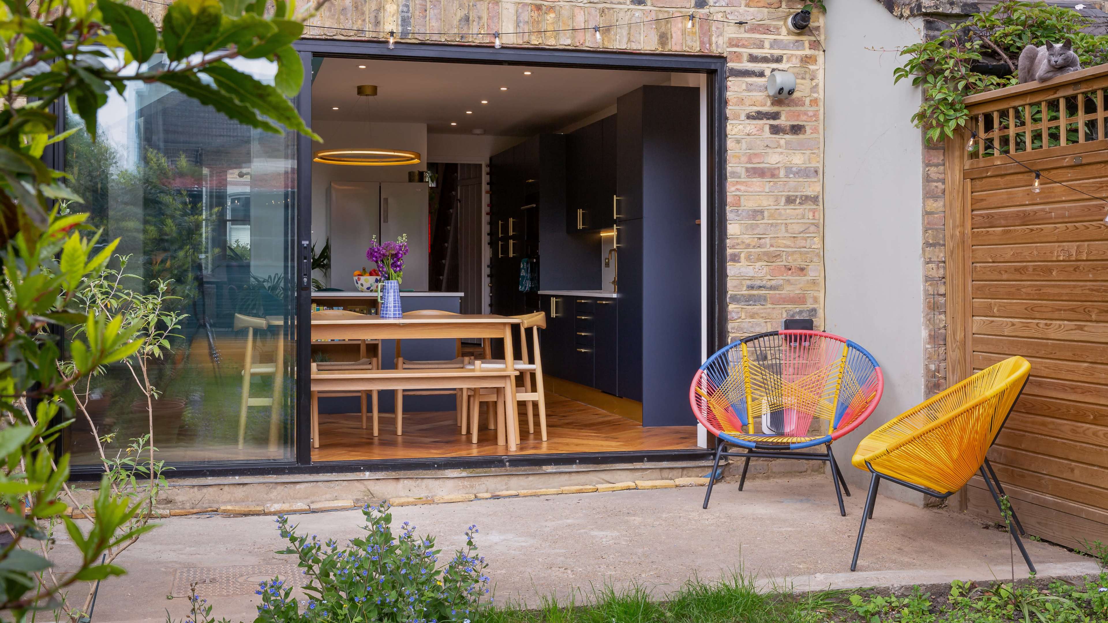 A modern kitchen and dining area with a wooden table and colorful chairs opens up to a garden featuring plants and a wooden fence.
