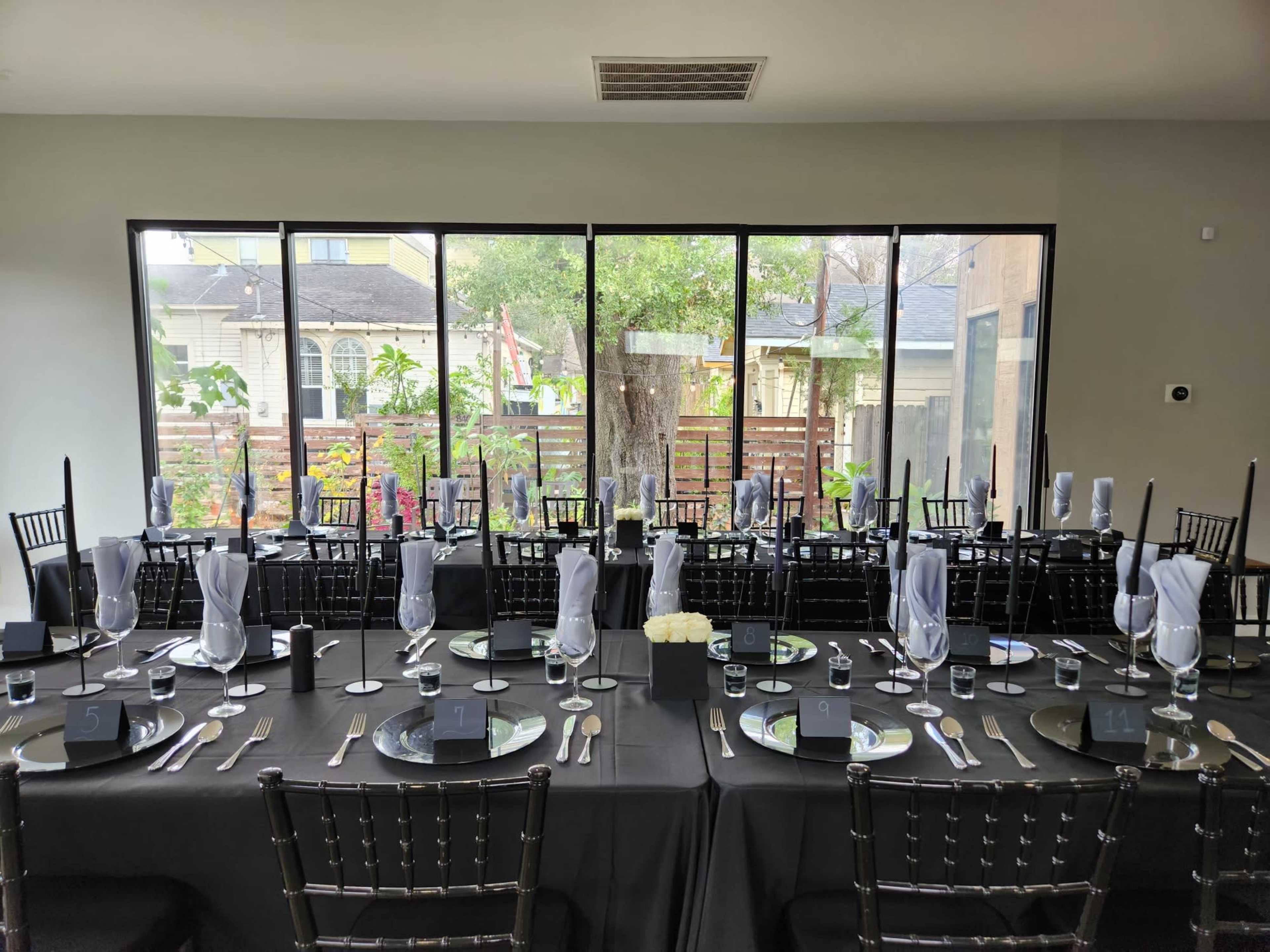 A long dining table is set with black tablecloths, silver cutlery, glassware, and white napkins, facing large windows overlooking a garden.