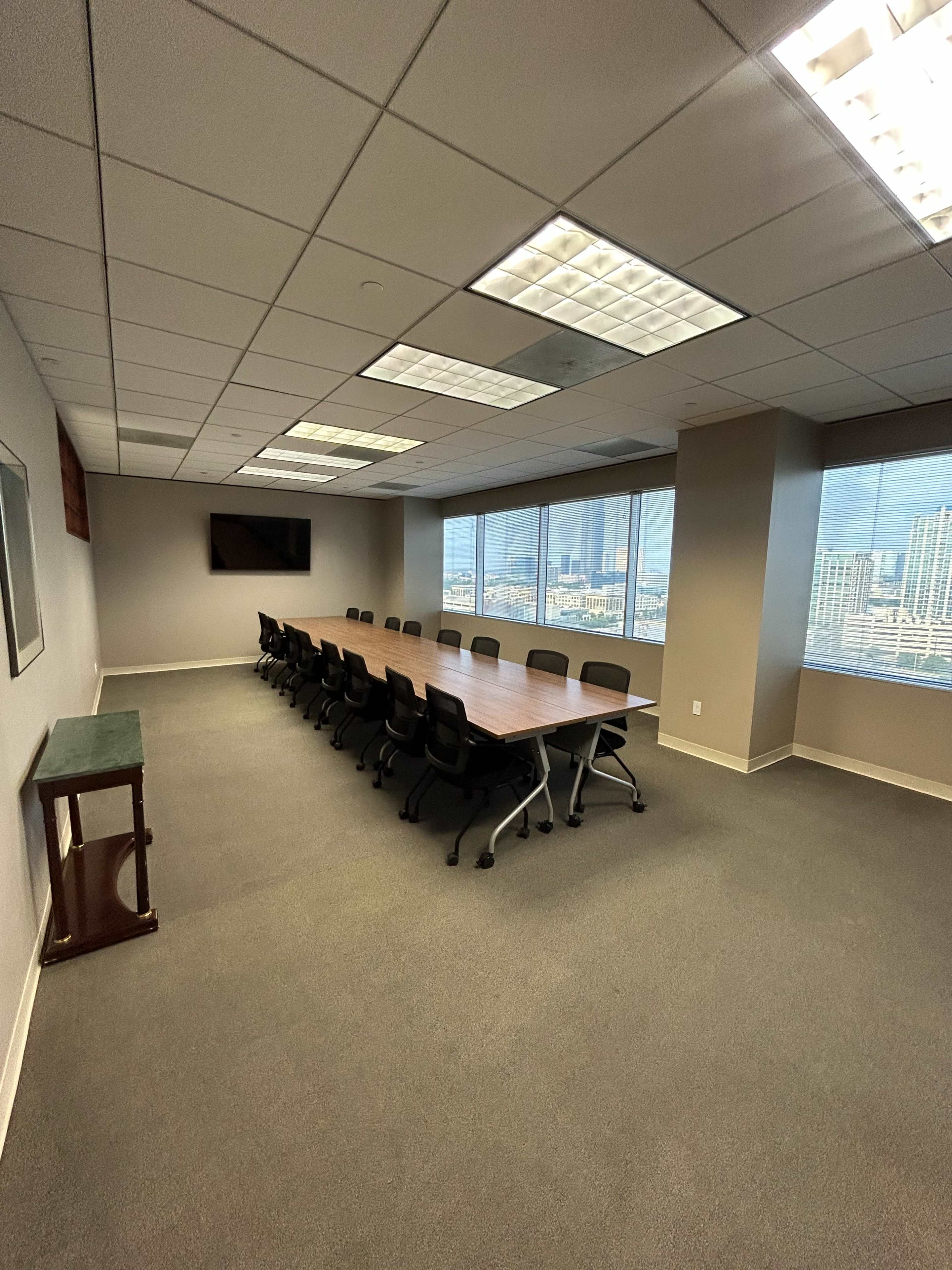 A large conference room features a long wooden table surrounded by black chairs, with windows offering a view of a city skyline.