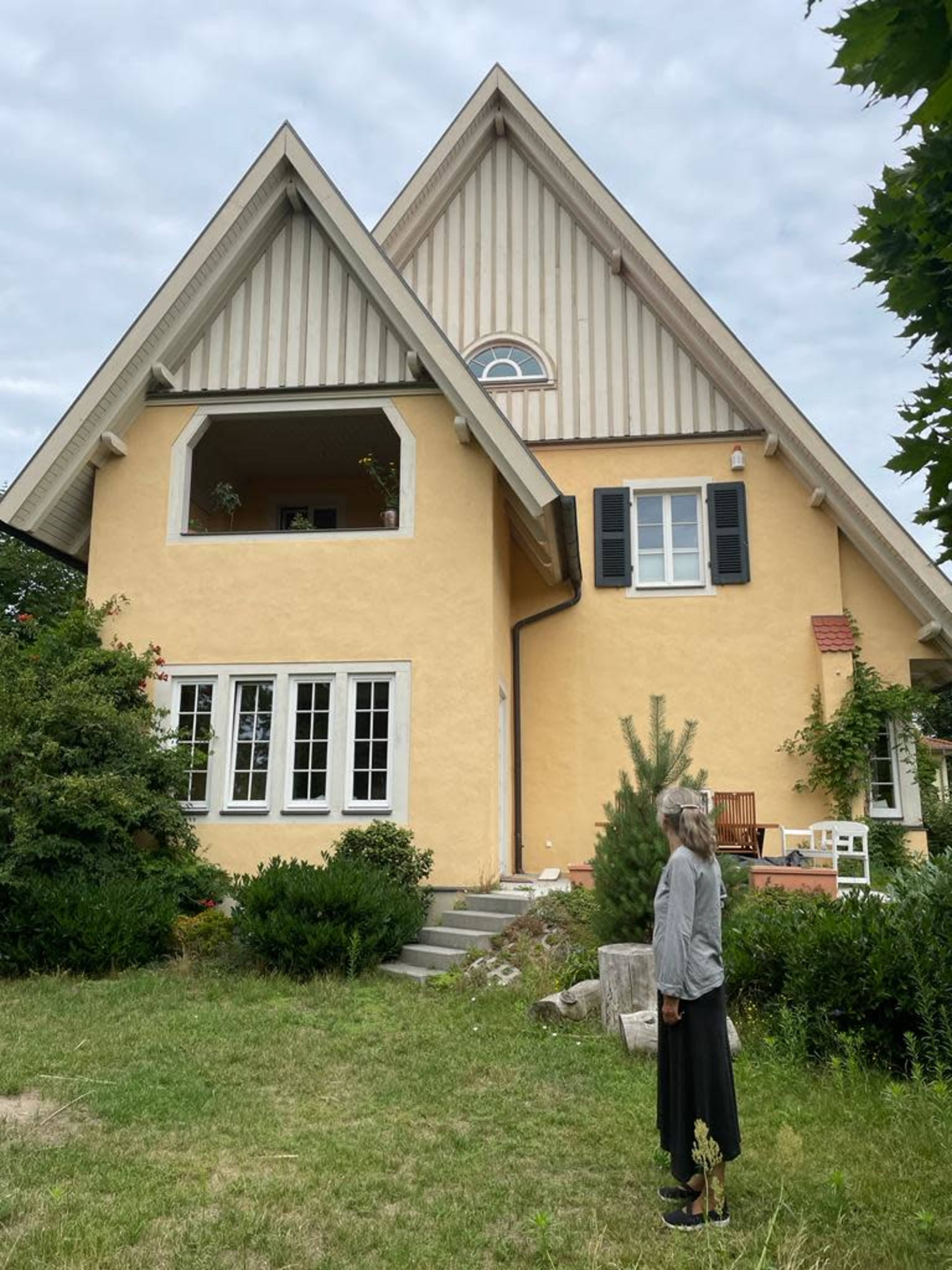 A woman stands in front of a large, yellow, two-story house with a peaked roof and front balcony, surrounded by greenery.