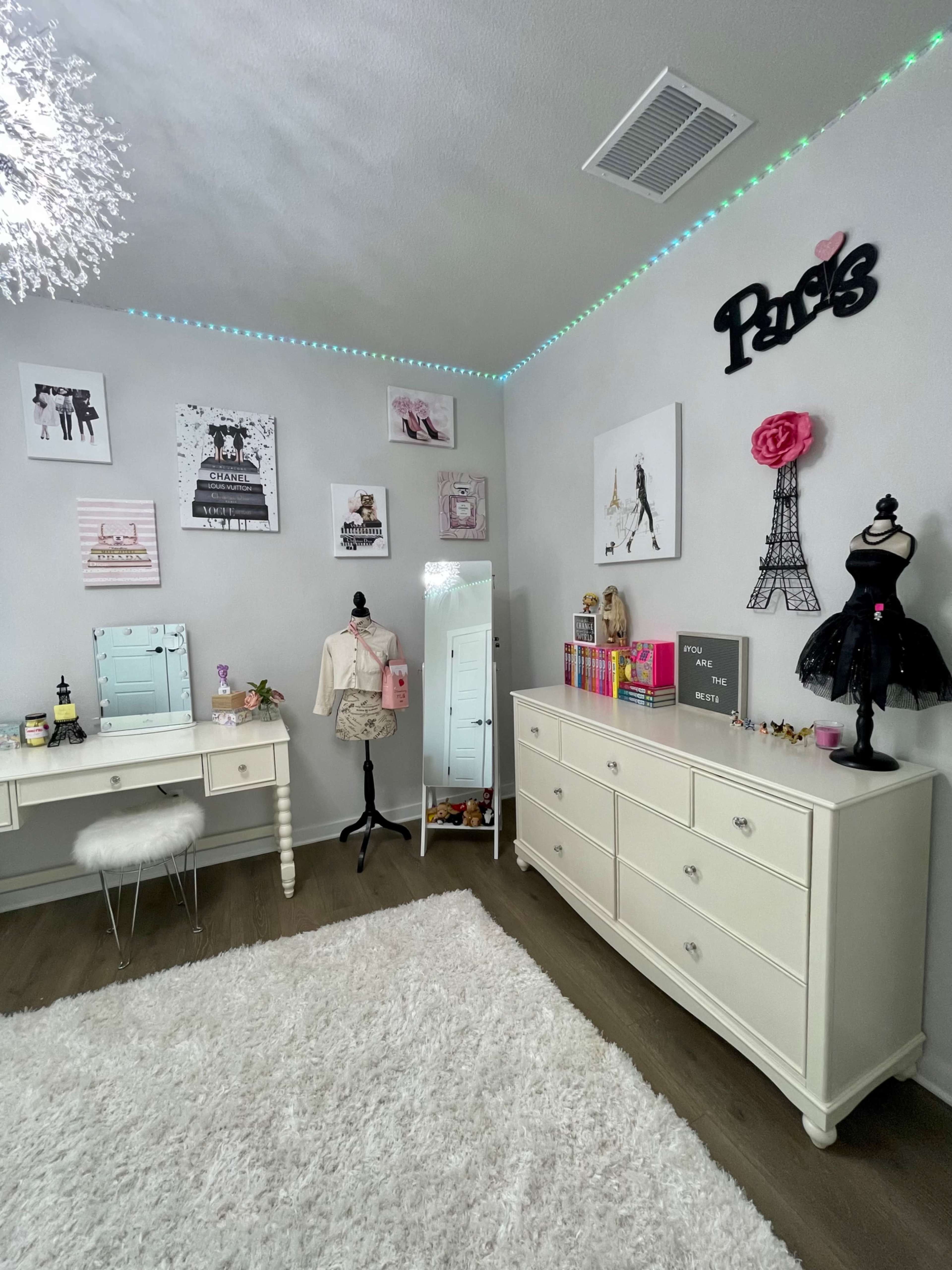 The image shows a neatly arranged bedroom featuring a white dresser, a vanity with a mirror, and elegant decor on the walls, including fashion-themed artwork and a mannequin dressed in a black outfit.