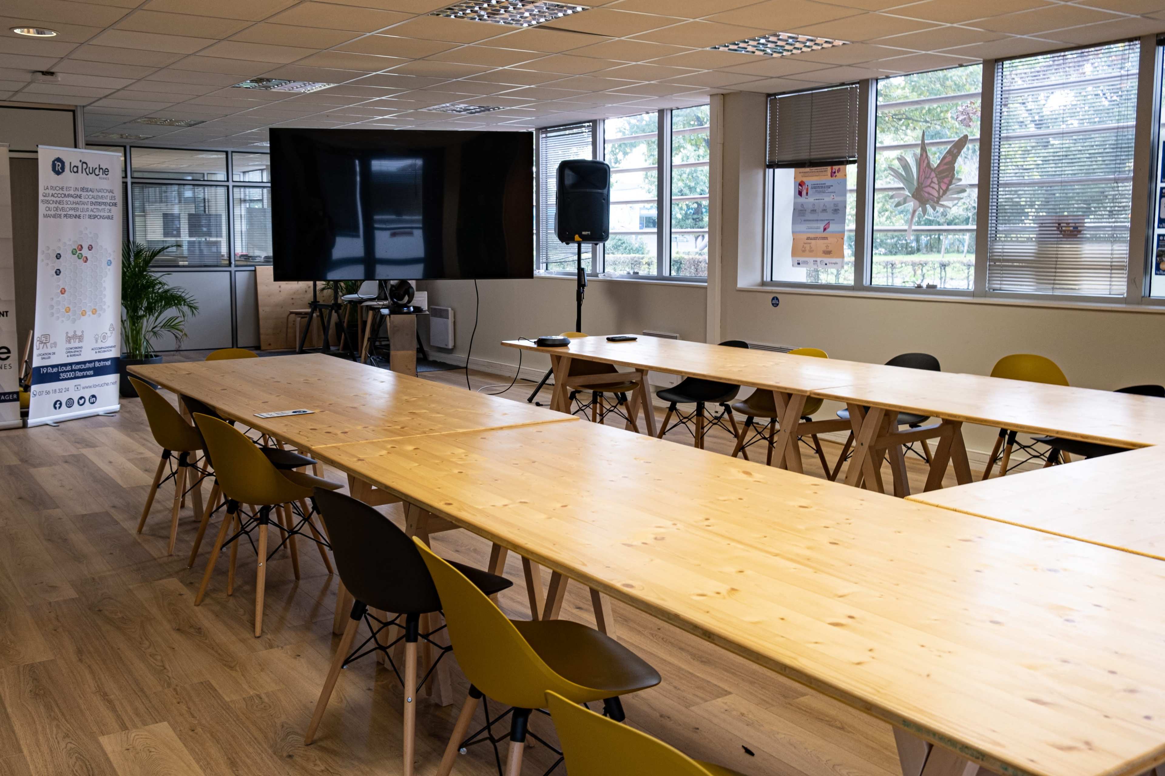 A large meeting room features several long wooden tables arranged in a U-shape, surrounded by chairs, with a television screen and a speaker in the corner.