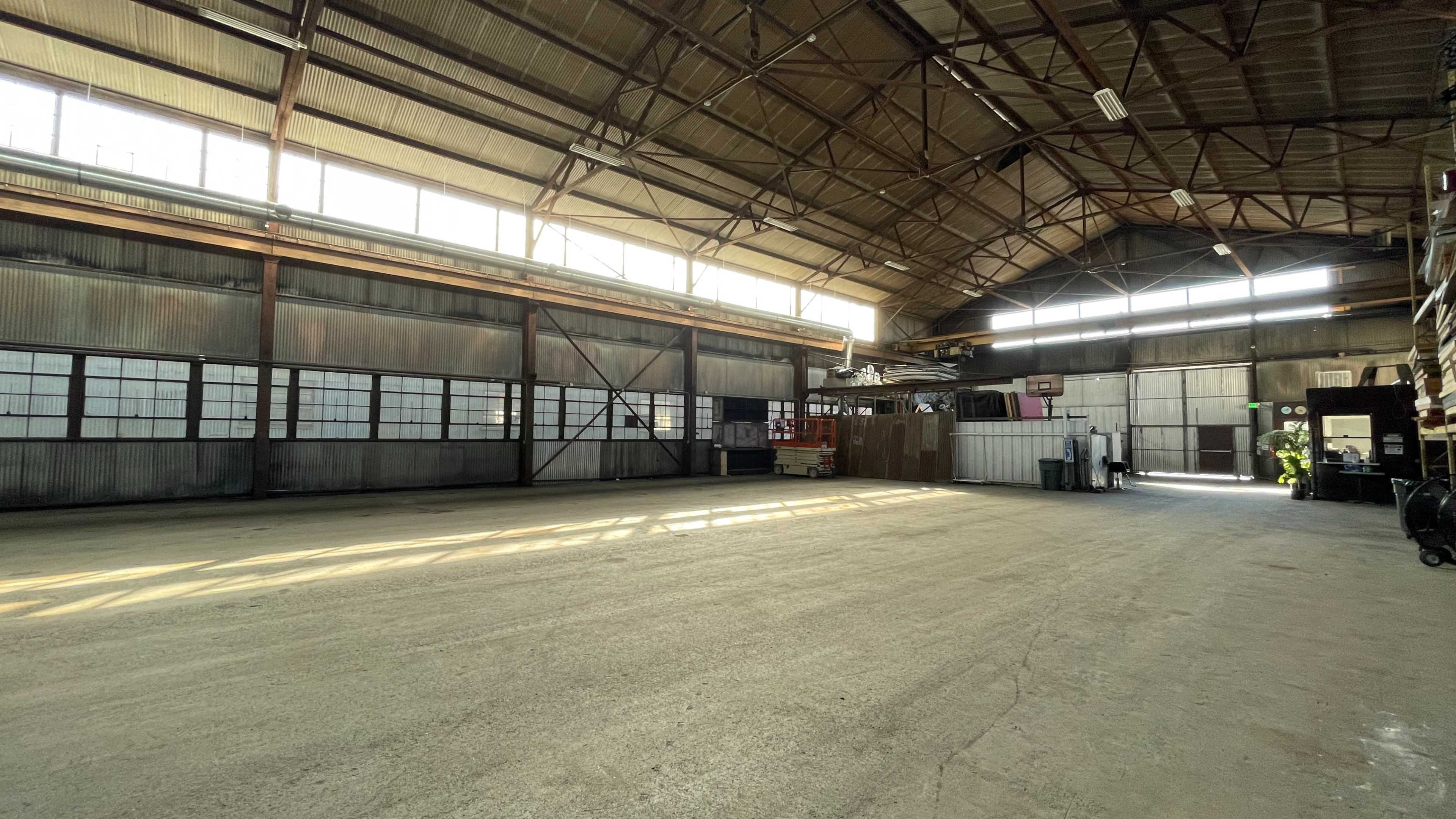 The image shows a spacious, industrial warehouse with large windows allowing natural light to enter, revealing a concrete floor and metal walls.