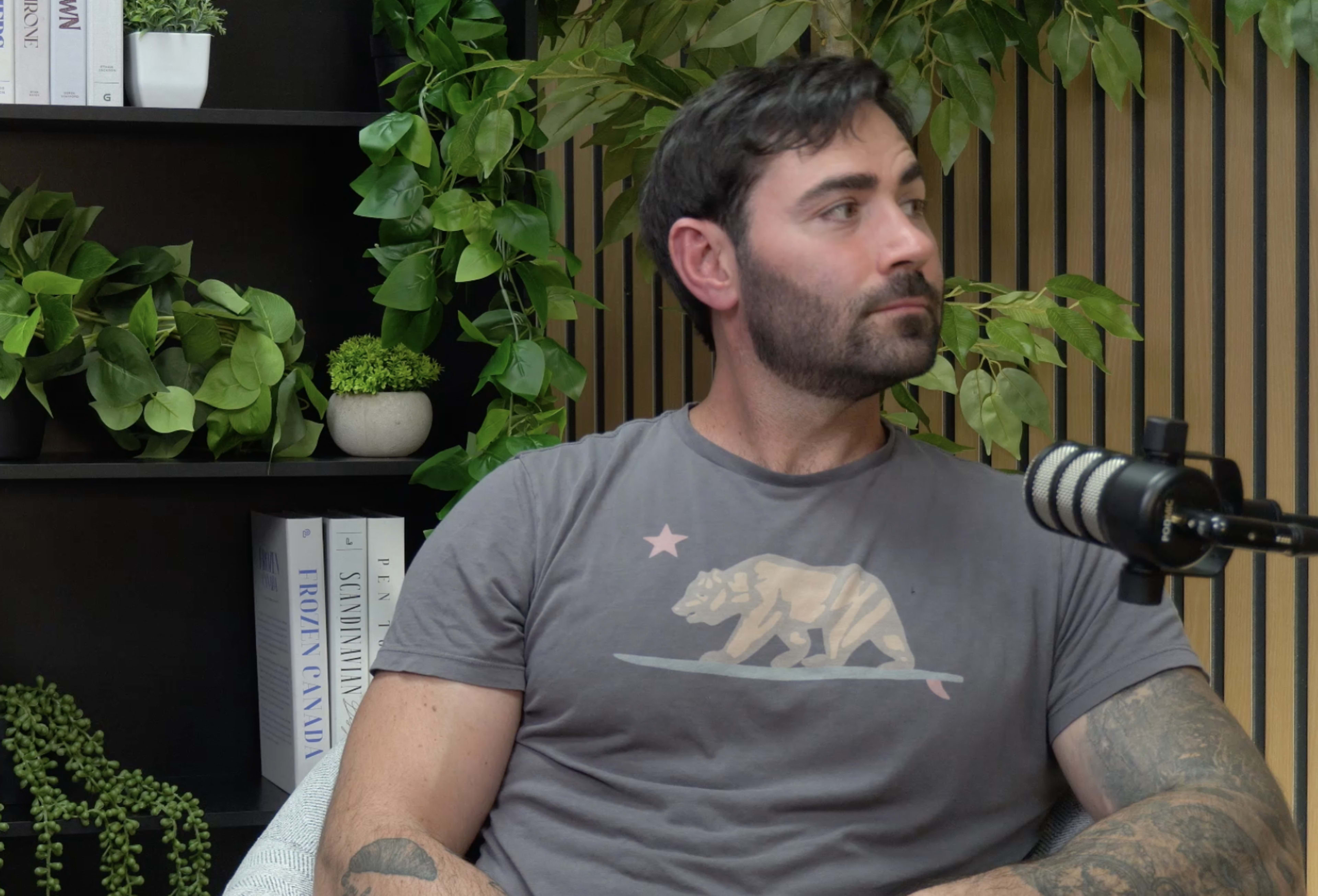 A man with a beard wearing a gray T-shirt sits next to a microphone against a backdrop of plants and shelves.