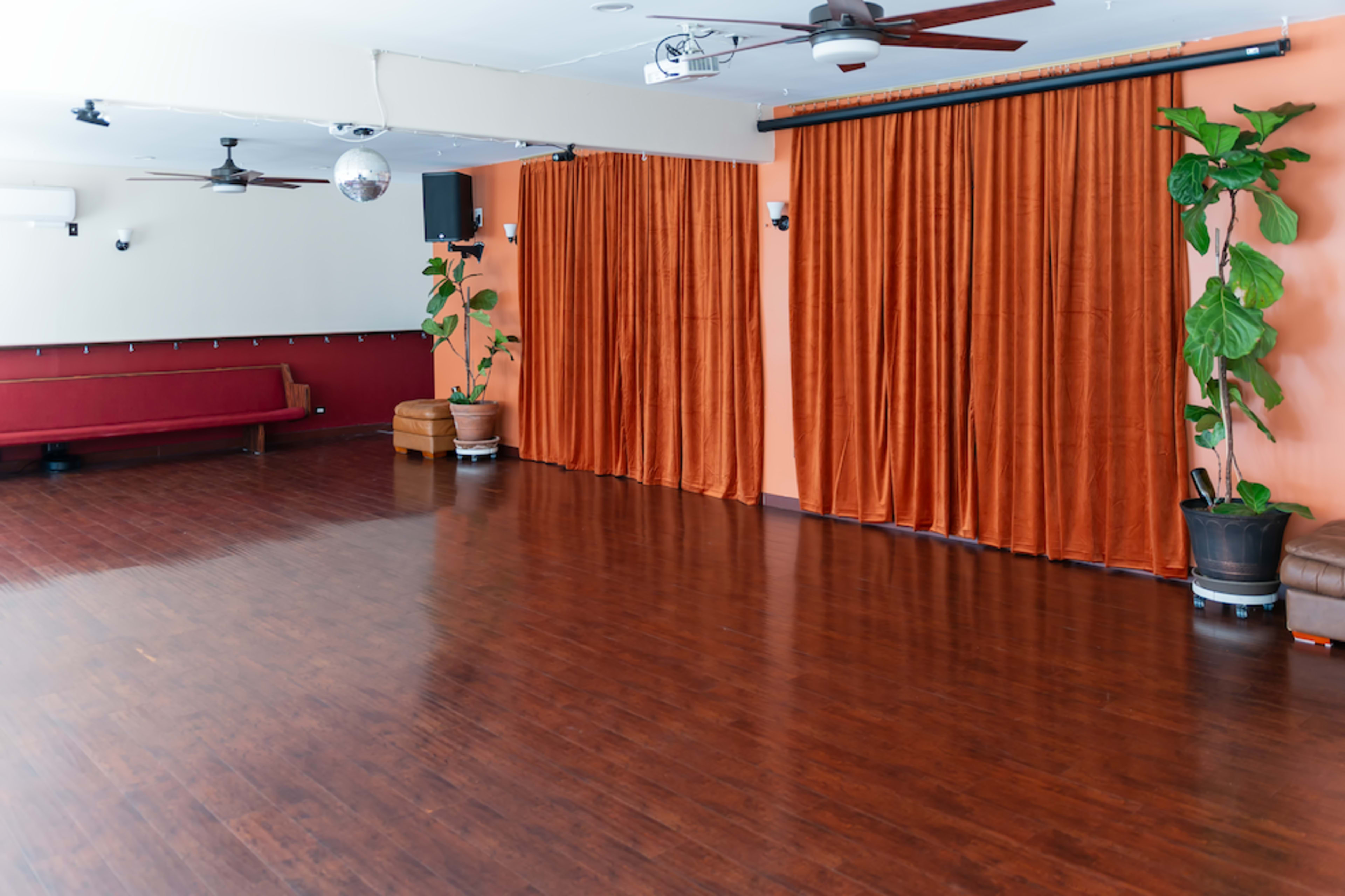 The image shows a spacious room with wooden flooring, orange curtains, two potted plants, and a seating area along one wall.