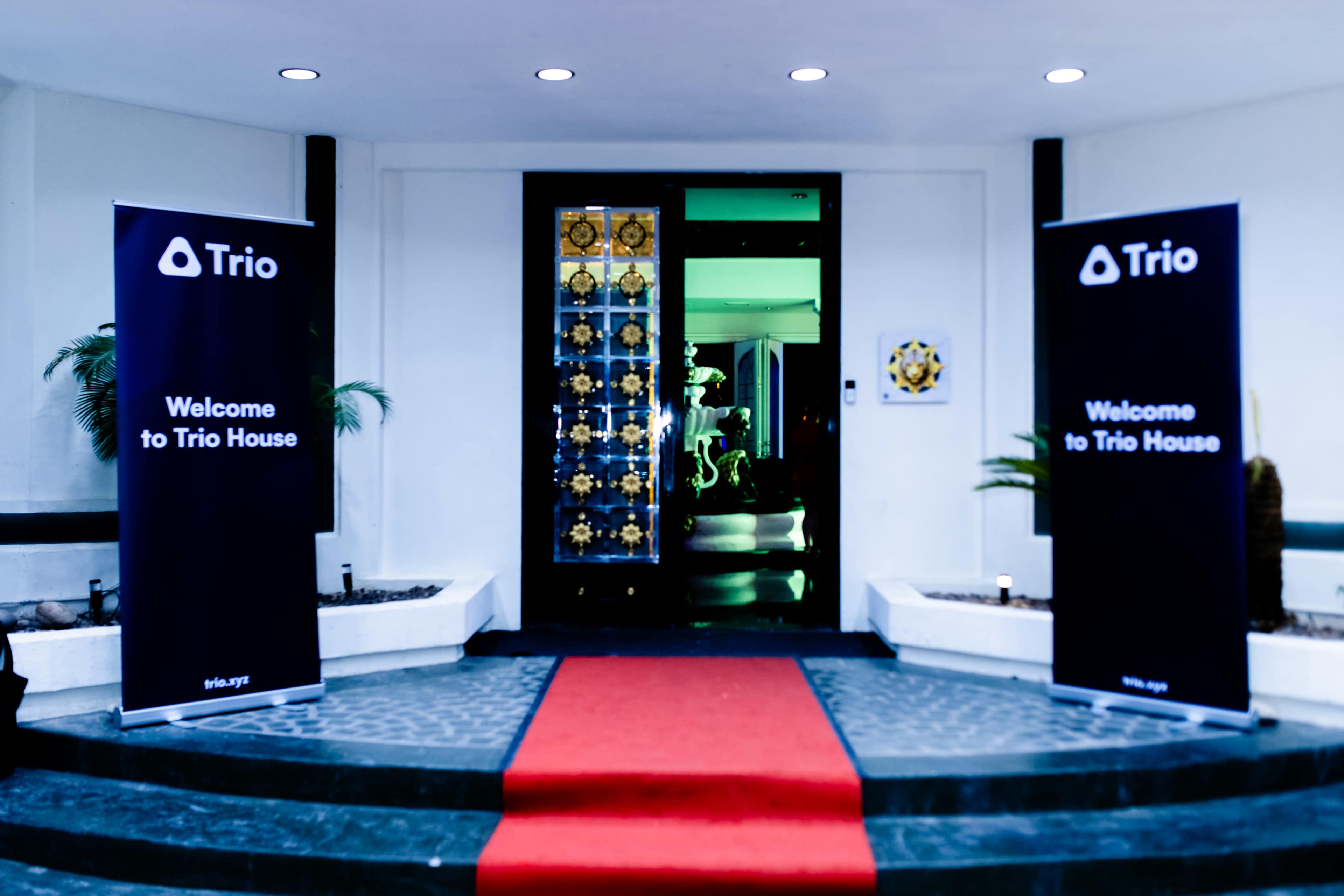 The image shows a red carpet leading to an entrance adorned with two banners welcoming guests to "Trio House" and featuring decorative doors behind them.