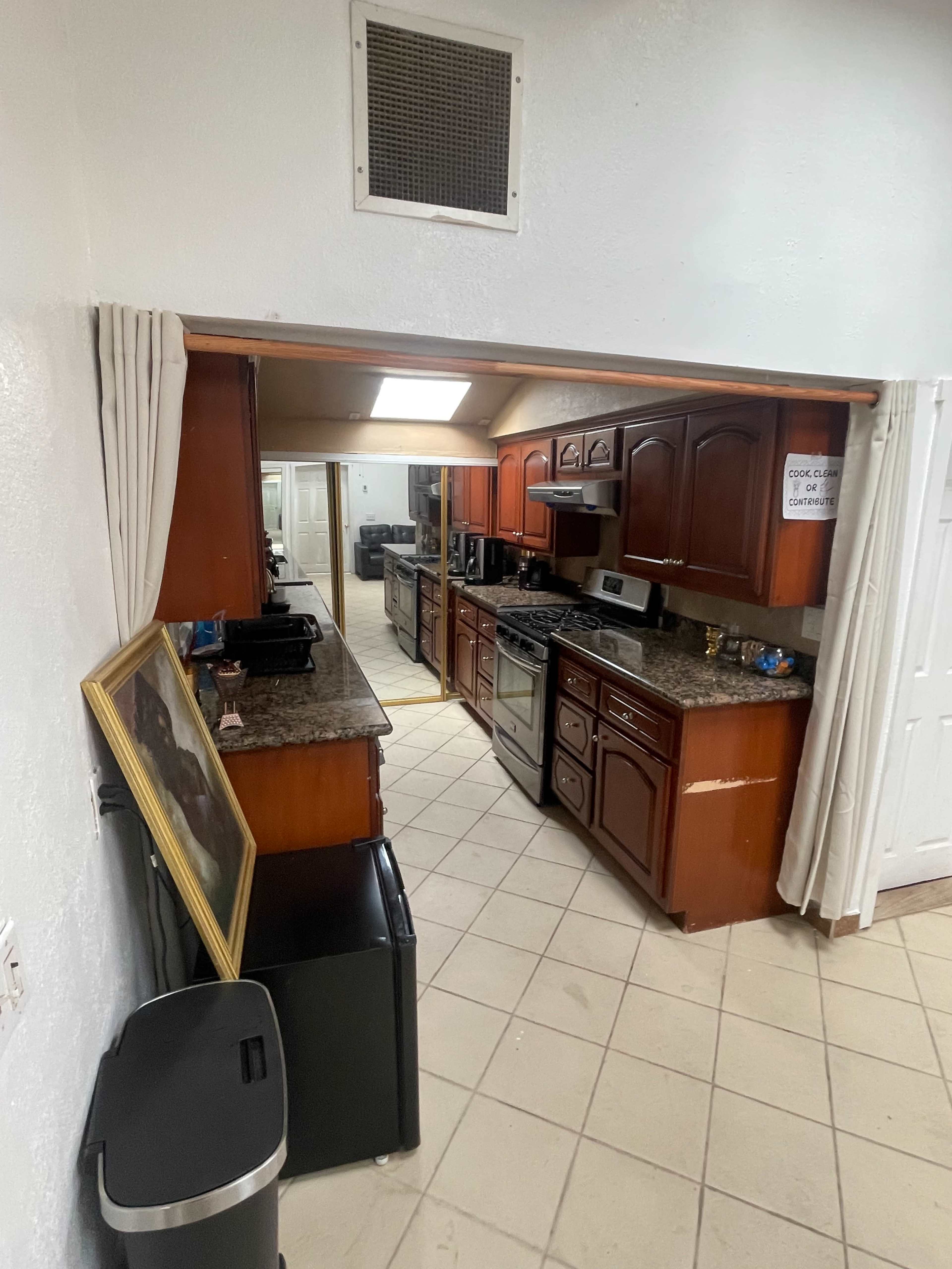 The image shows a kitchen with wooden cabinets, a granite countertop, and various appliances, seen from an adjacent room through an open doorway.