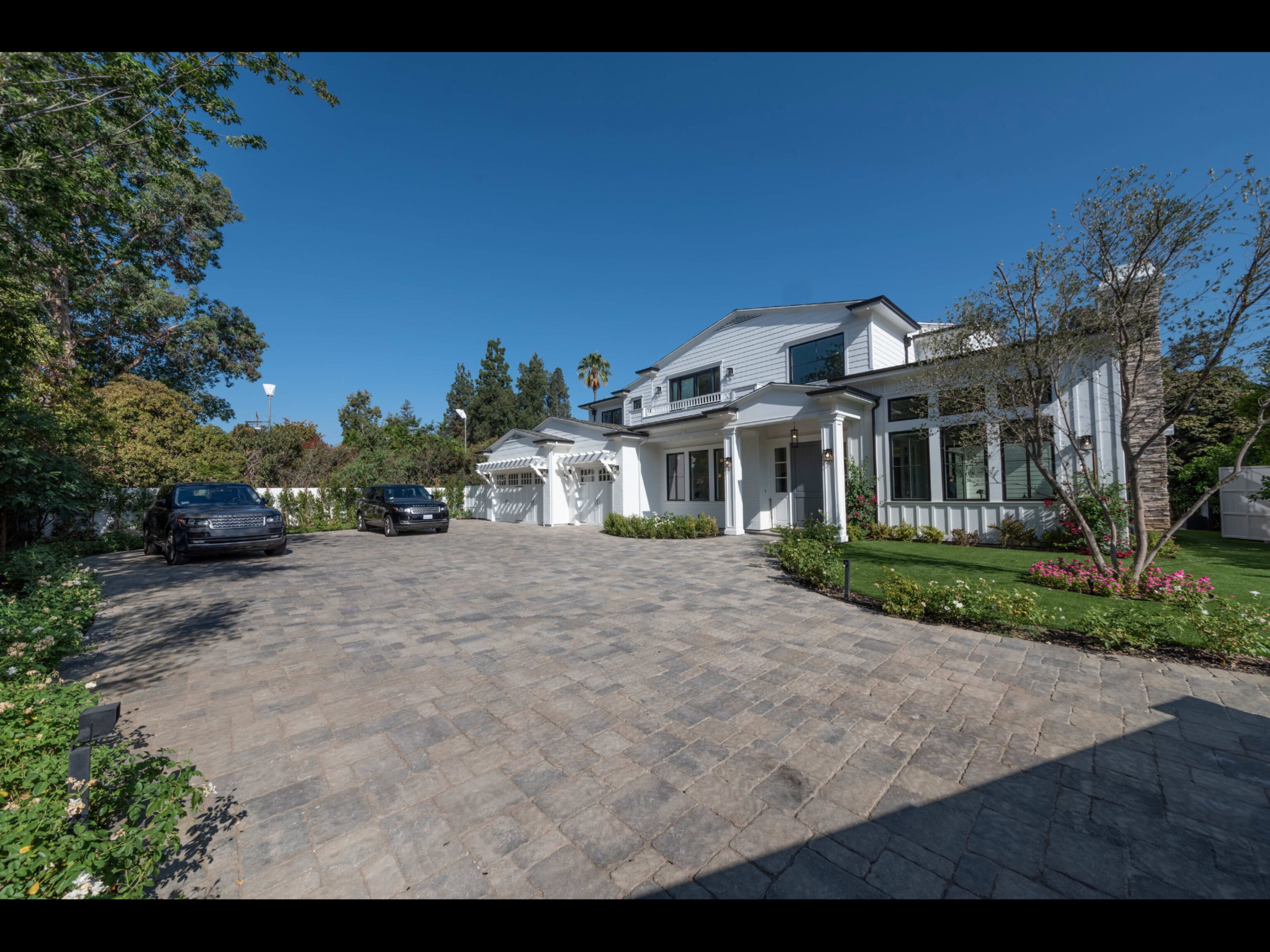 The image shows a large modern house with a wide driveway made of pavers, surrounded by lush greenery and landscaping under a clear sky.