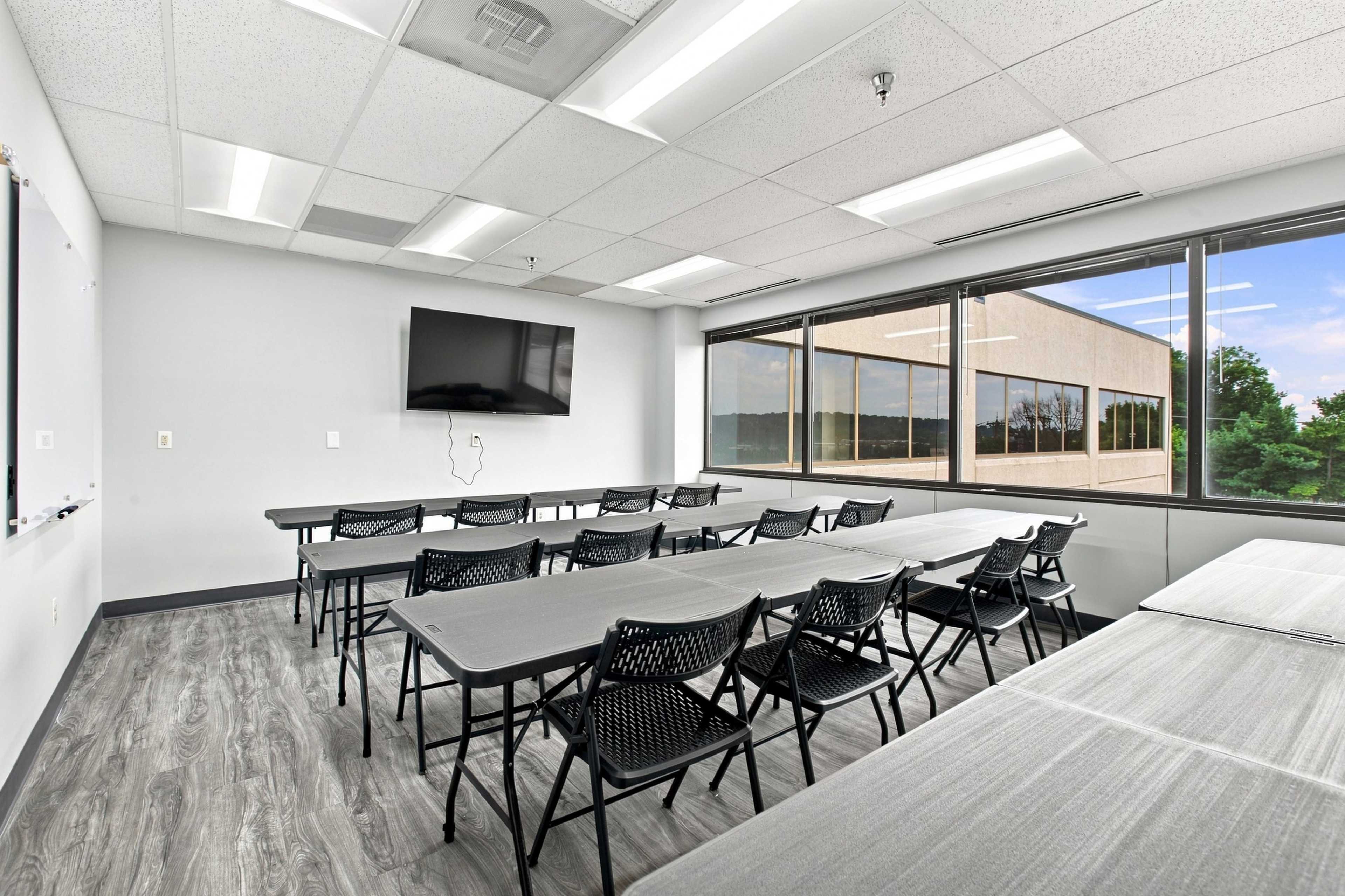 A bright classroom features several rows of folding tables and chairs facing a large wall-mounted television and a window showcasing an outdoor view.