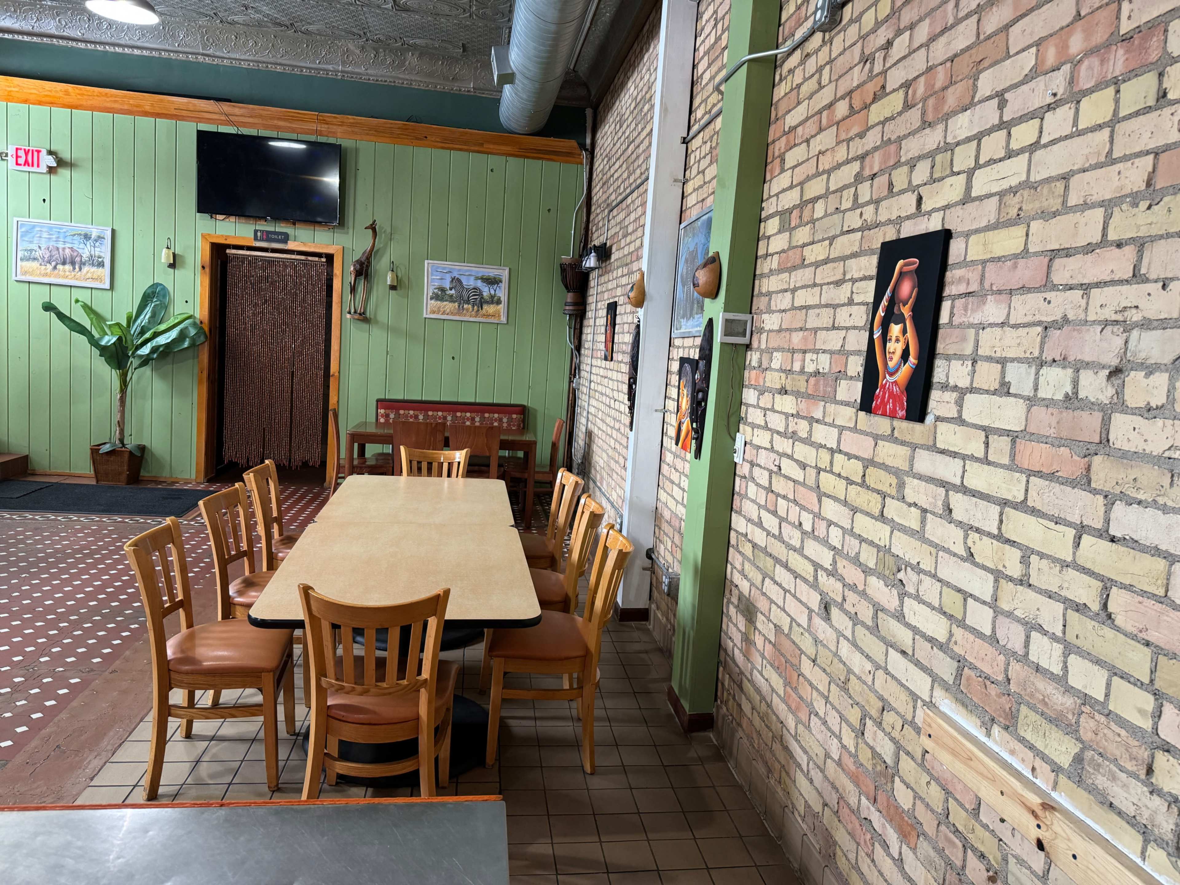 A long table with wooden chairs is set against a brick wall in a restaurant featuring green paneling and framed artwork.